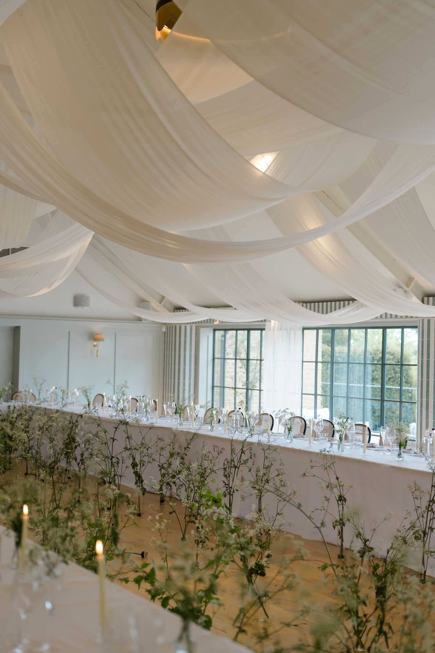 Elegant wedding reception hall with white draped ceiling, long tables, floral greenery centerpieces, and tall windows.