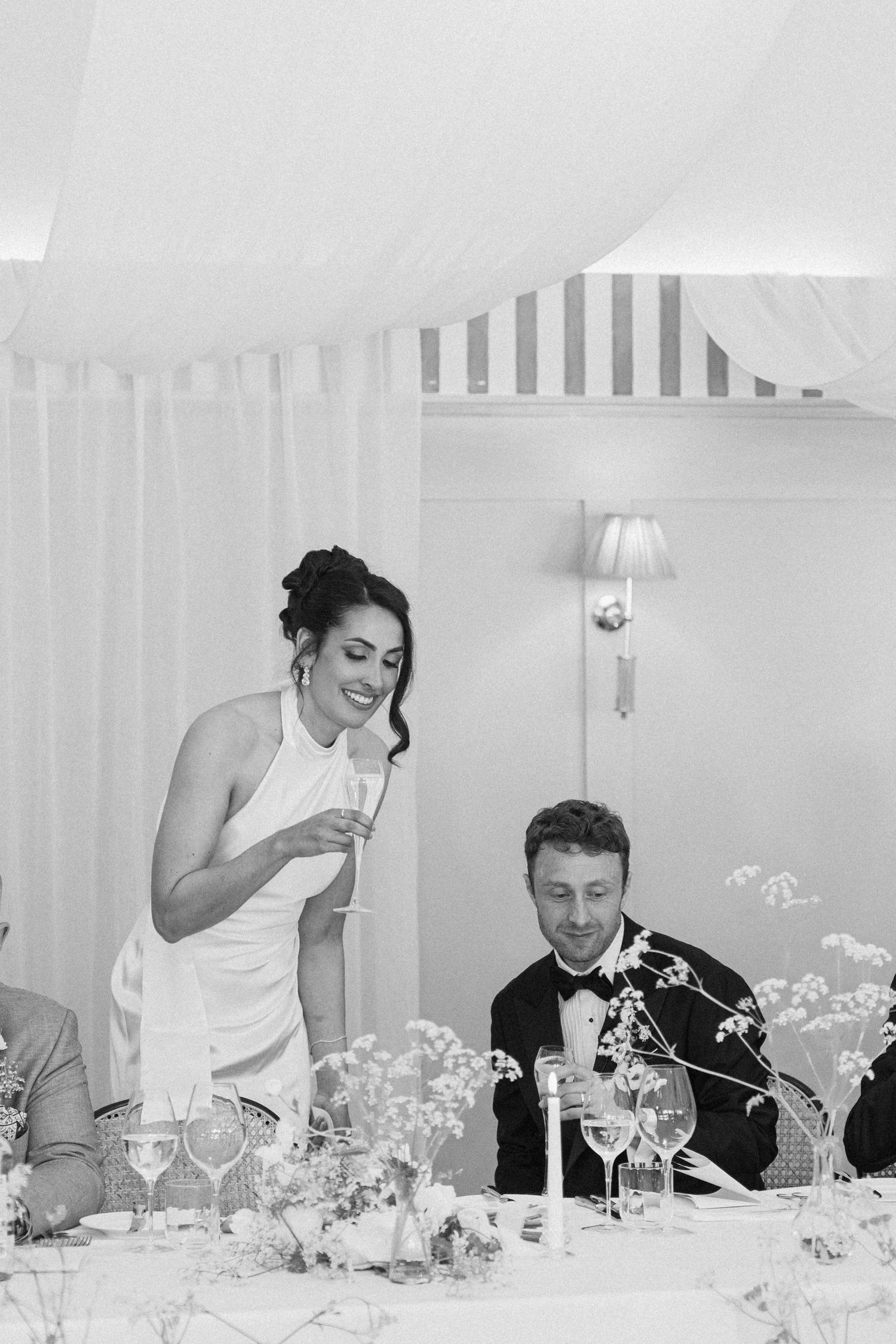 Black and white photo of a bride smiling while giving a wedding speech holding a champagne glass.
