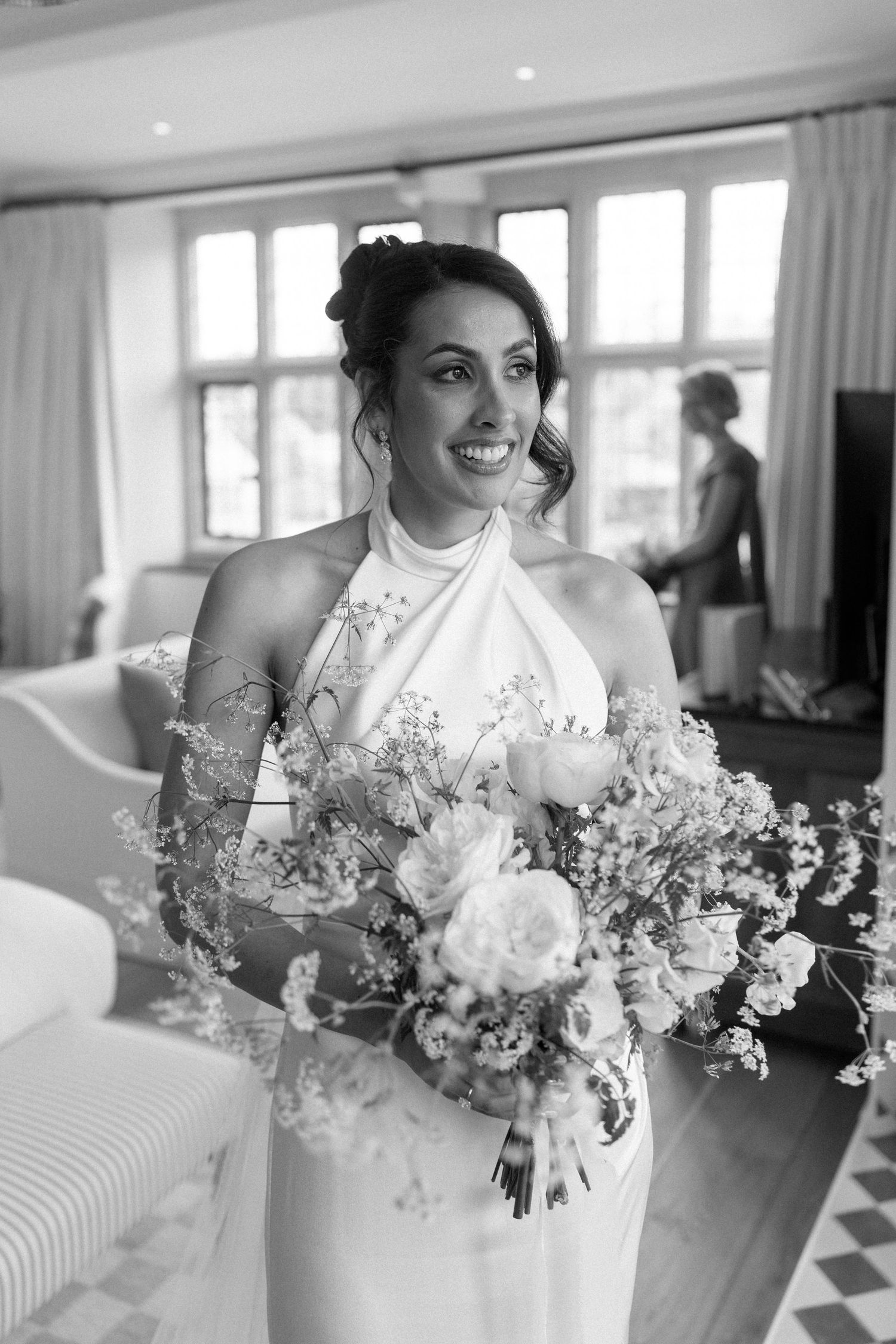 Smiling bride in a halter neck wedding dress holds a white floral bouquet in a bright indoor setting.