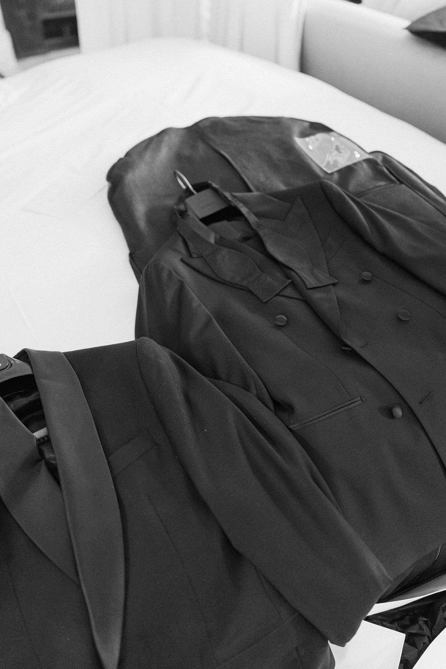 A black and white close-up of a dark double-breasted suit jacket laid flat on a white bed before a wedding.