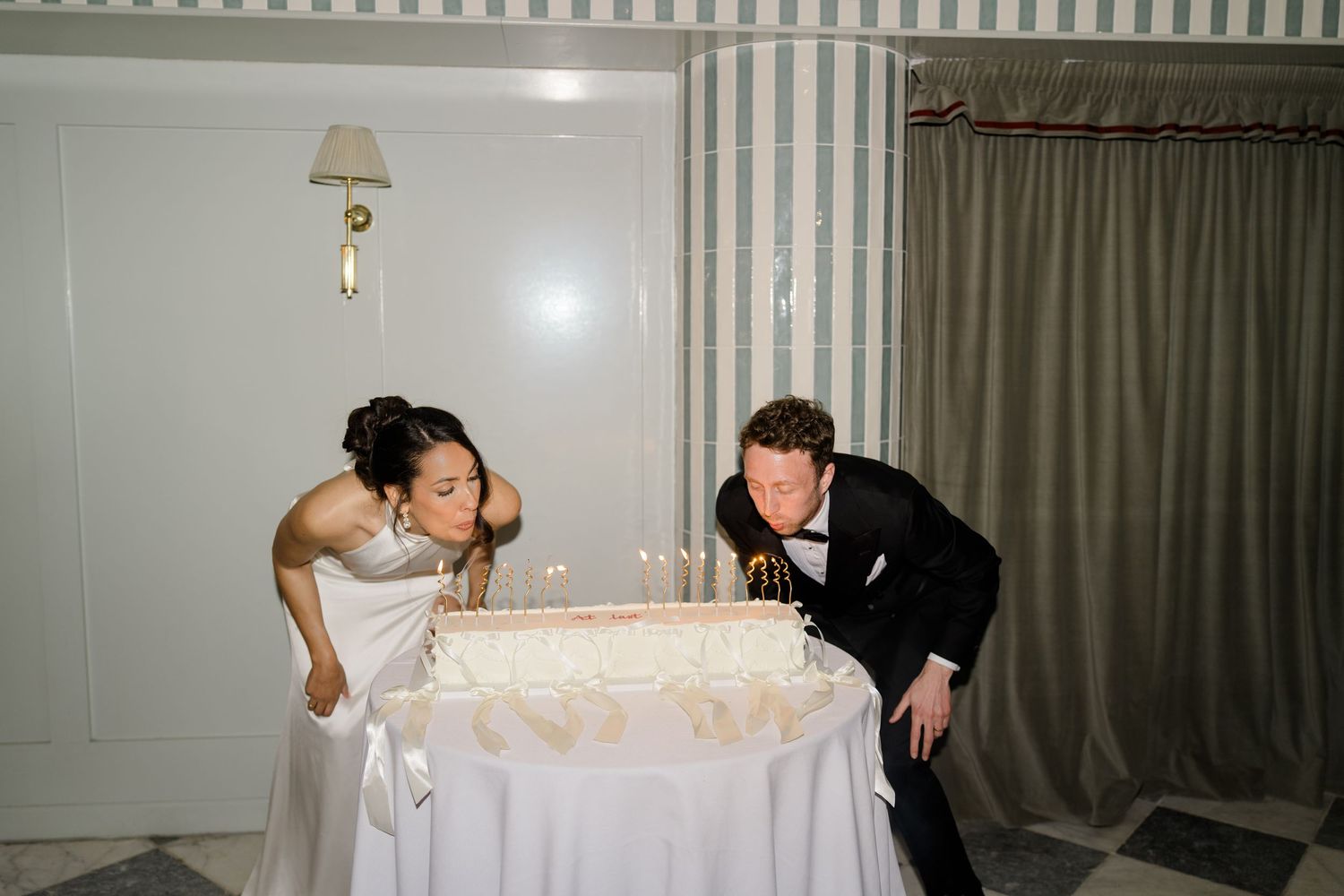 Bride and groom blow out candles on a white wedding cake in a striped reception room.
