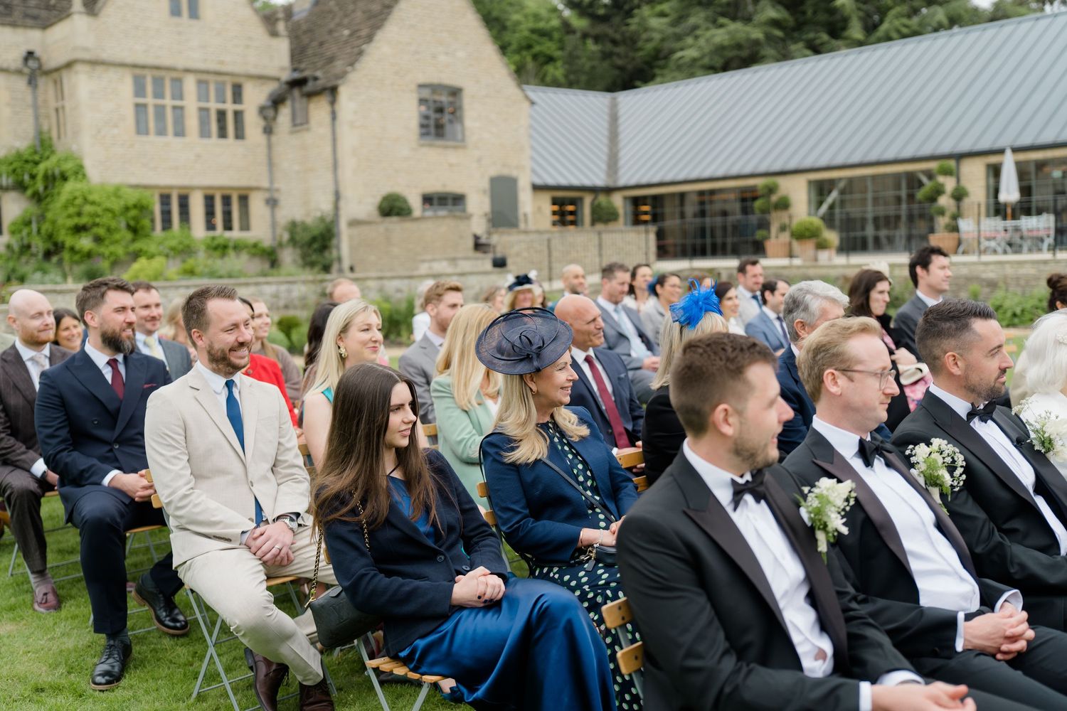 Wedding guests seated outdoors in front of a beautiful stone manor house during a garden ceremony.