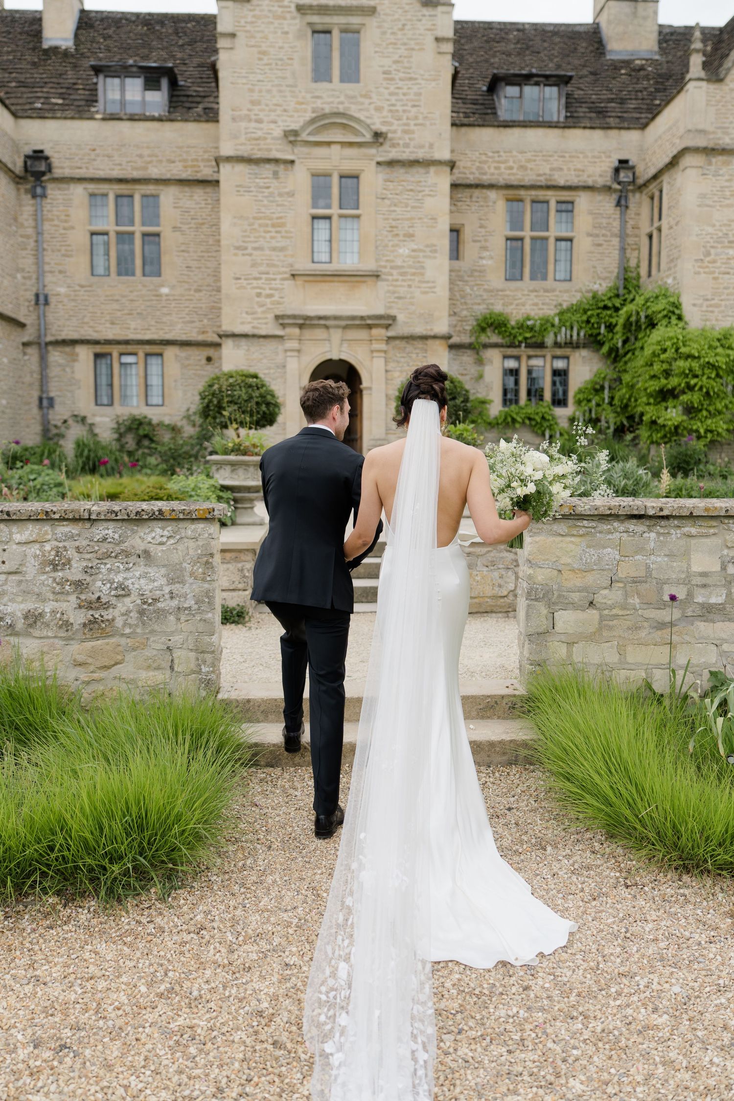 Bride and groom walking hand in hand toward a grand stone manor house, bride wearing a flowing veil.