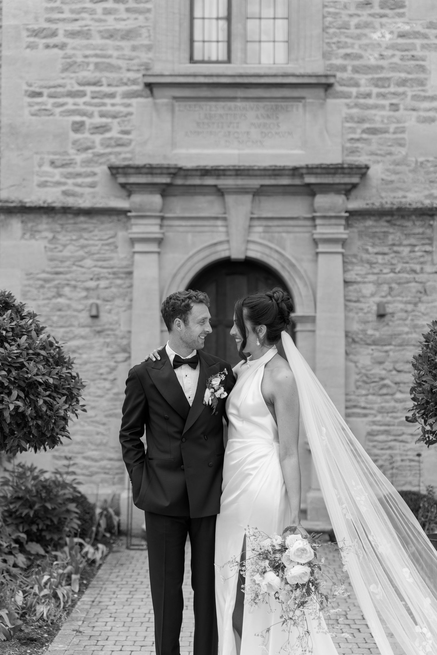 Bride and groom share an intimate glance outside a historic stone church in black and white.