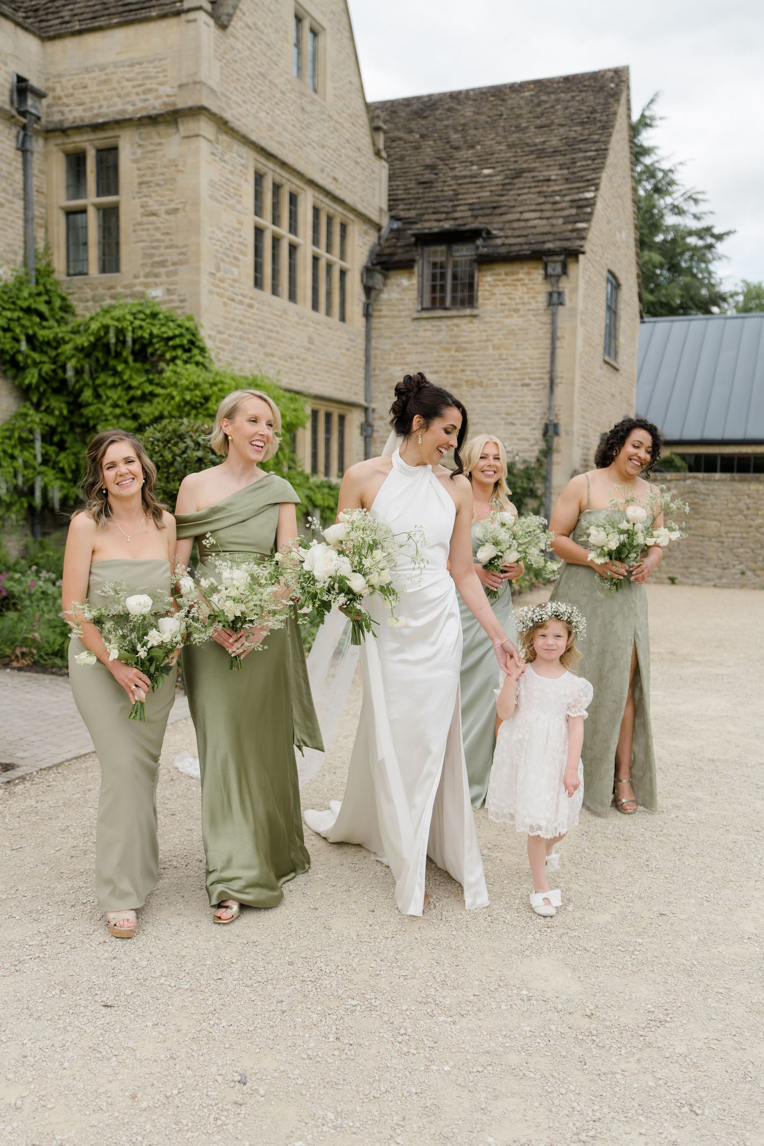 Bride in sleek satin gown walks with bridesmaids in sage green dresses and flower girl outside stone manor venue.