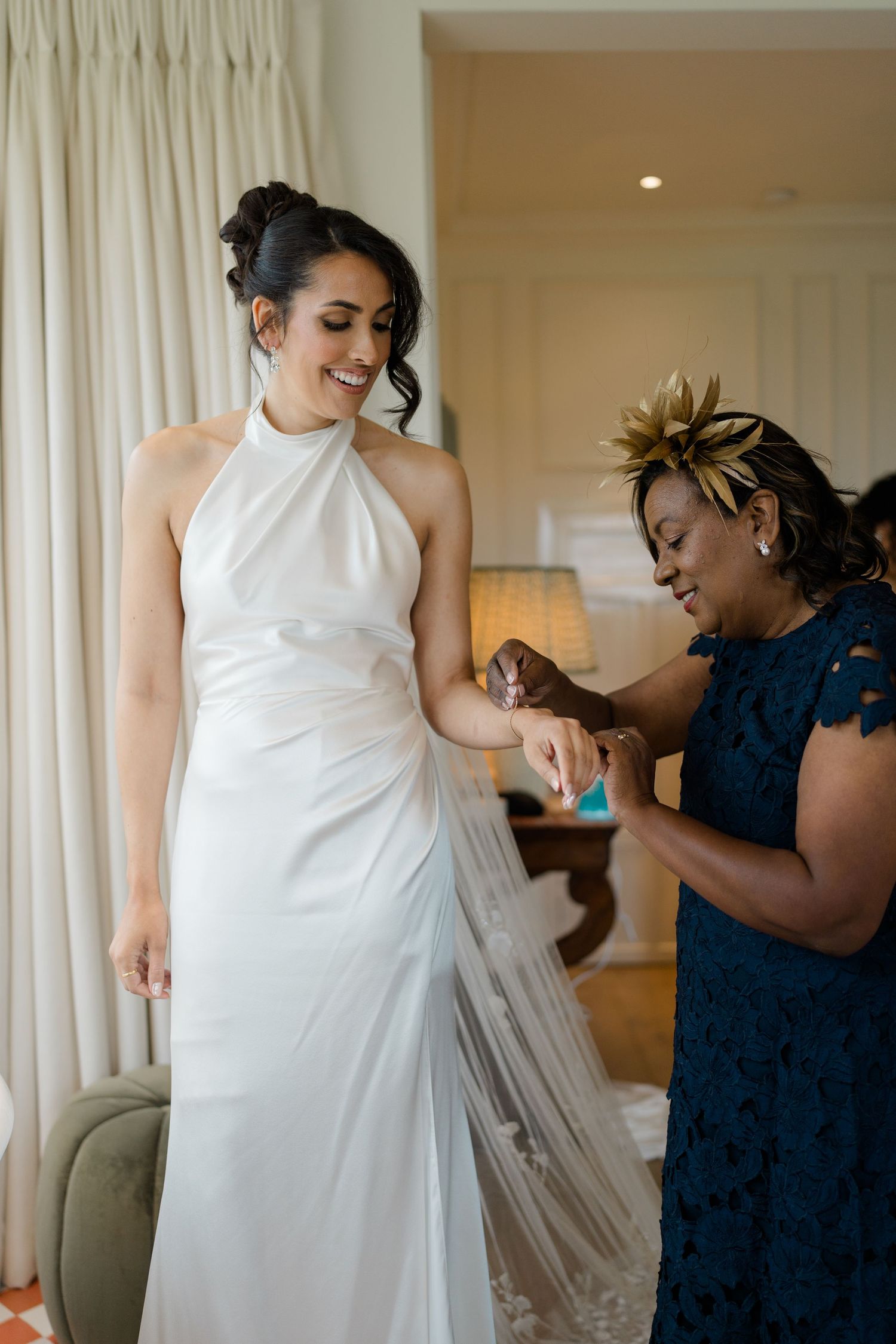 Bride in sleek halter neck wedding dress smiling as guest sprays perfume on her wrist.