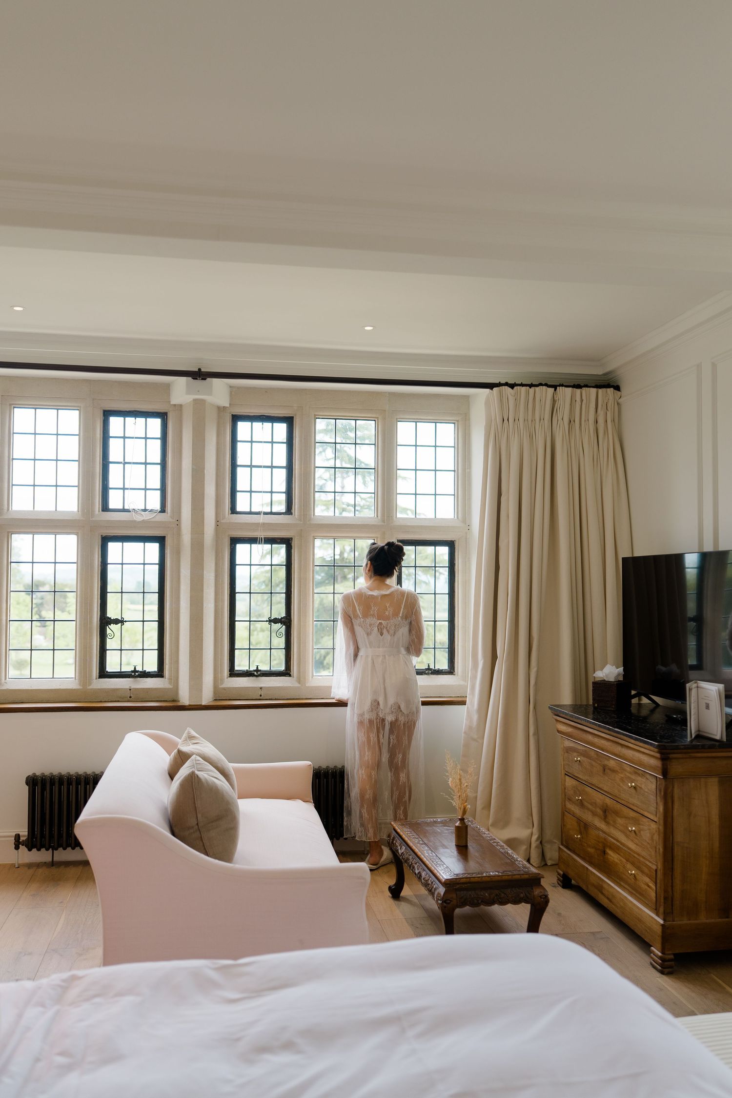 Bride in white lace robe standing at large leaded glass windows in a luxury manor bedroom.