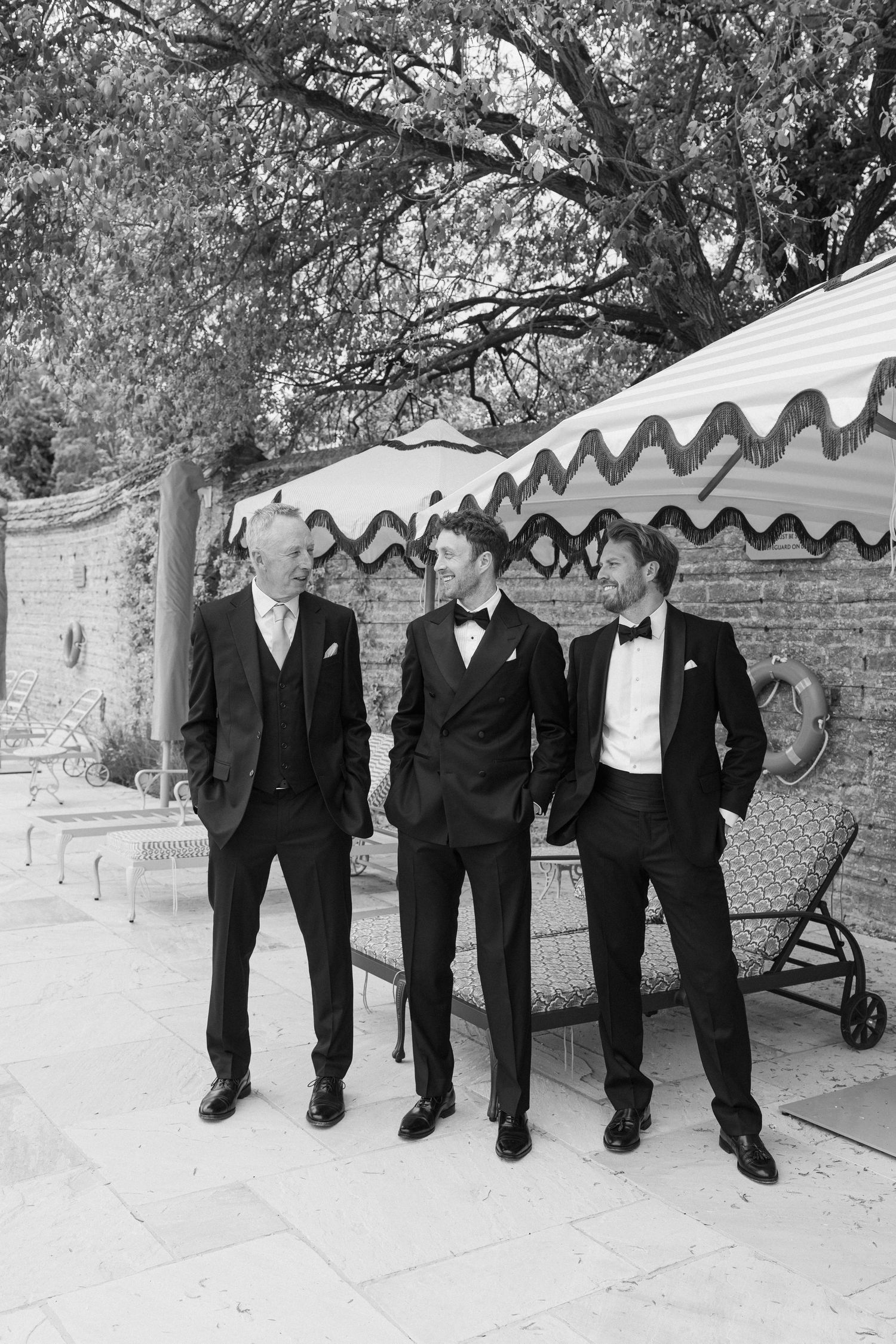 Black and white photo of groom and groomsmen in tuxedos laughing together beside a striped marquee.
