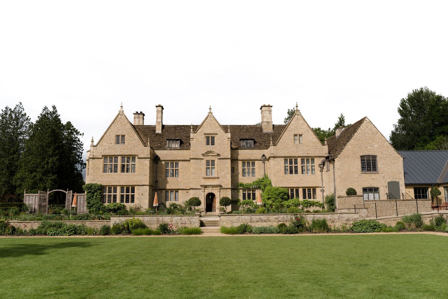A grand Cotswold stone manor house with manicured lawns and lush green gardens on an overcast day.