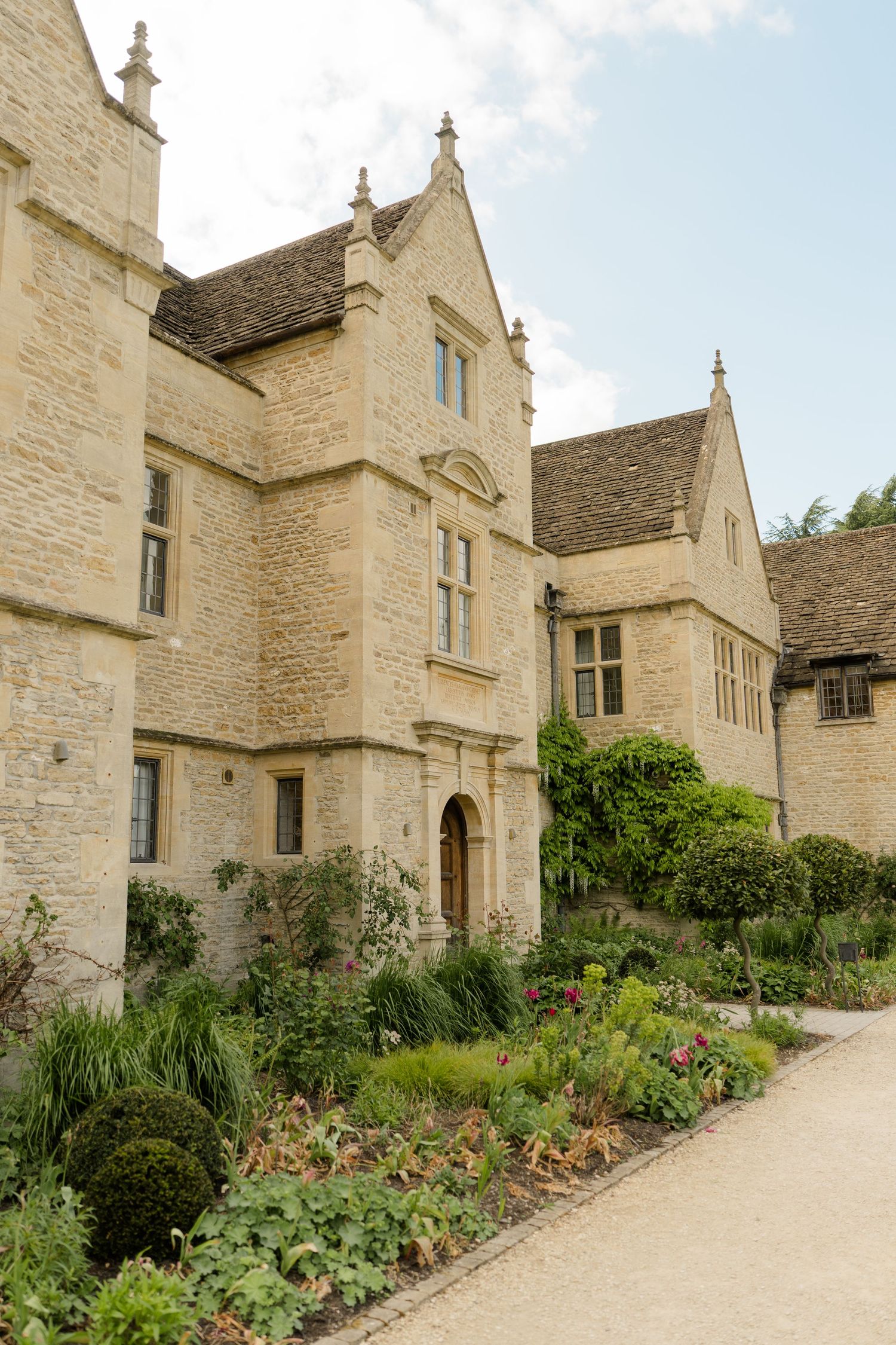 Historic Cotswold stone manor house with manicured garden borders and gravel pathway on a cloudy day.