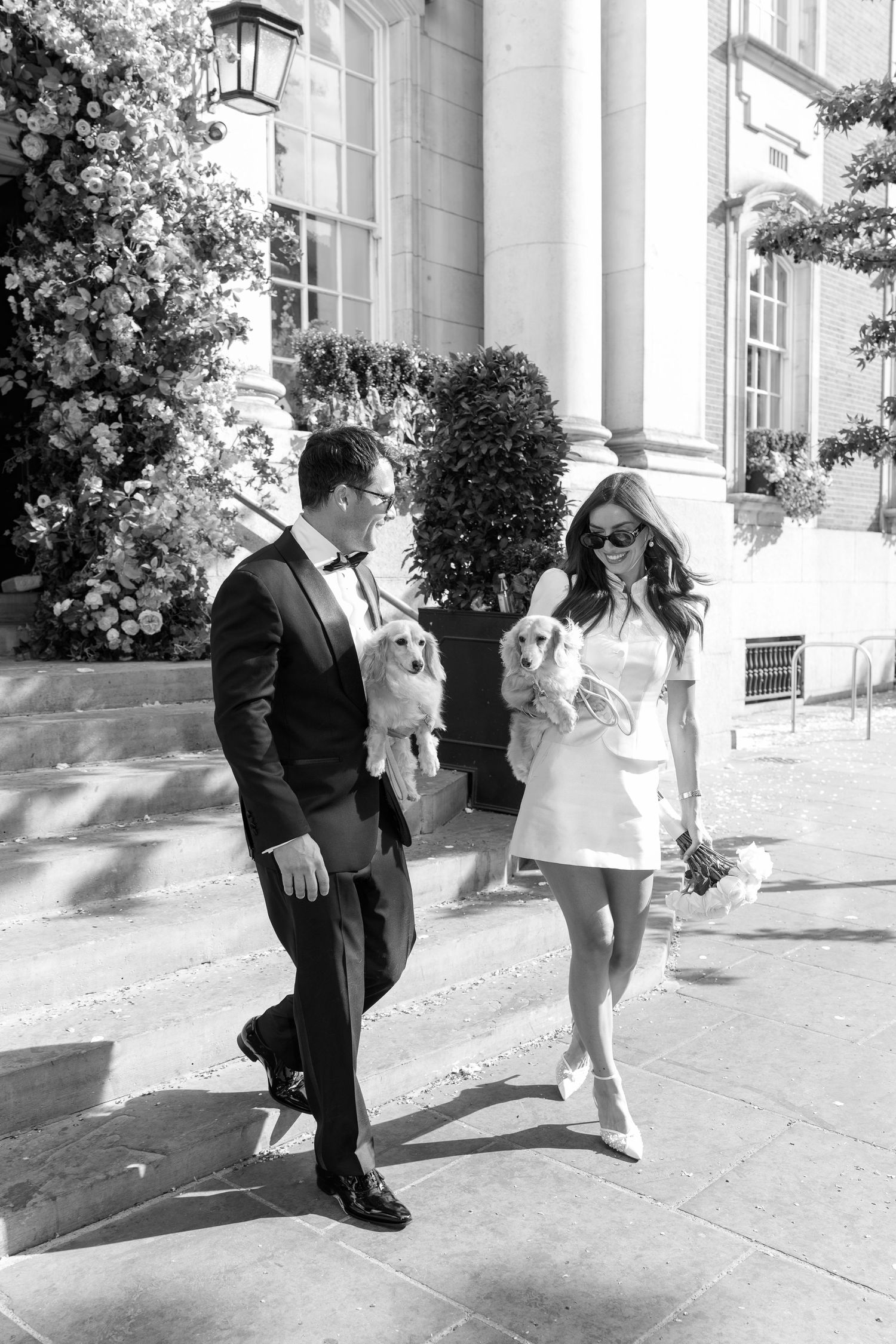 Black and white photo of bride and groom each holding a puppy outside an elegant building.