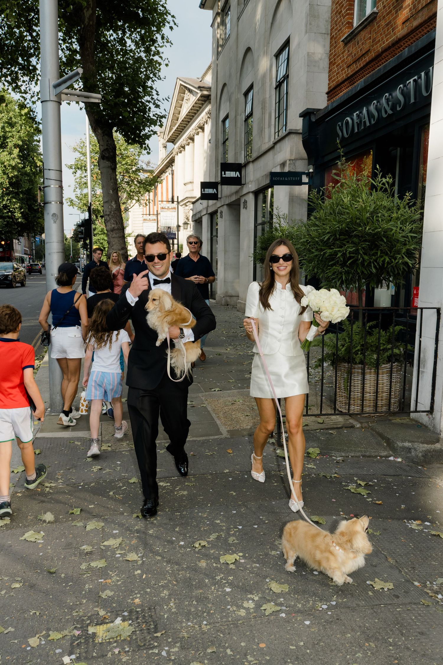 Bride in white mini dress holding bouquet walks dog with groom in black suit on sunny urban street.