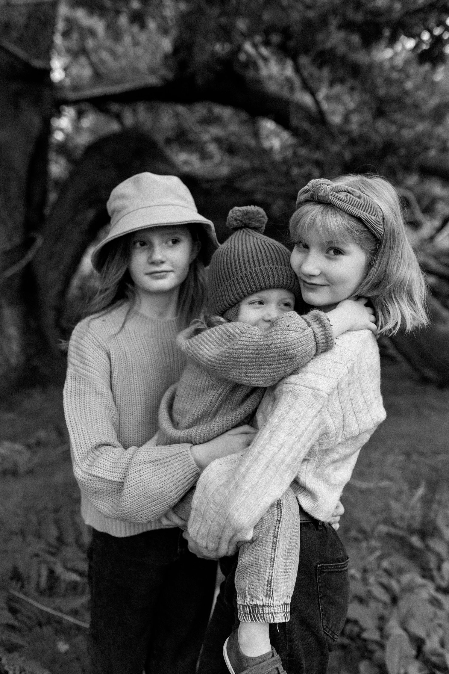 Three people in cozy sweaters share a warm embrace outdoors in black and white.
