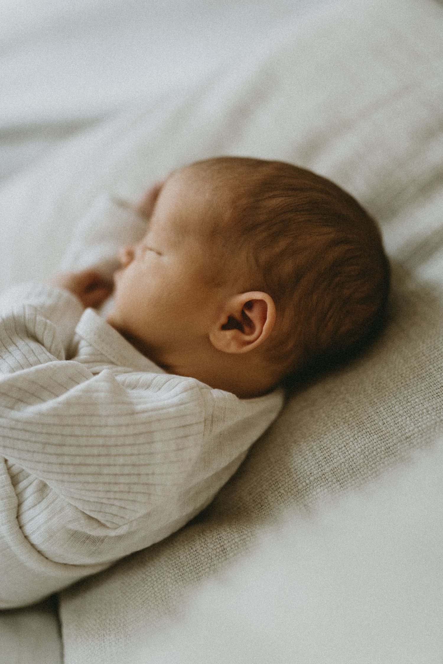 Newborn baby in white ribbed onesie sleeping on light colored bedding.