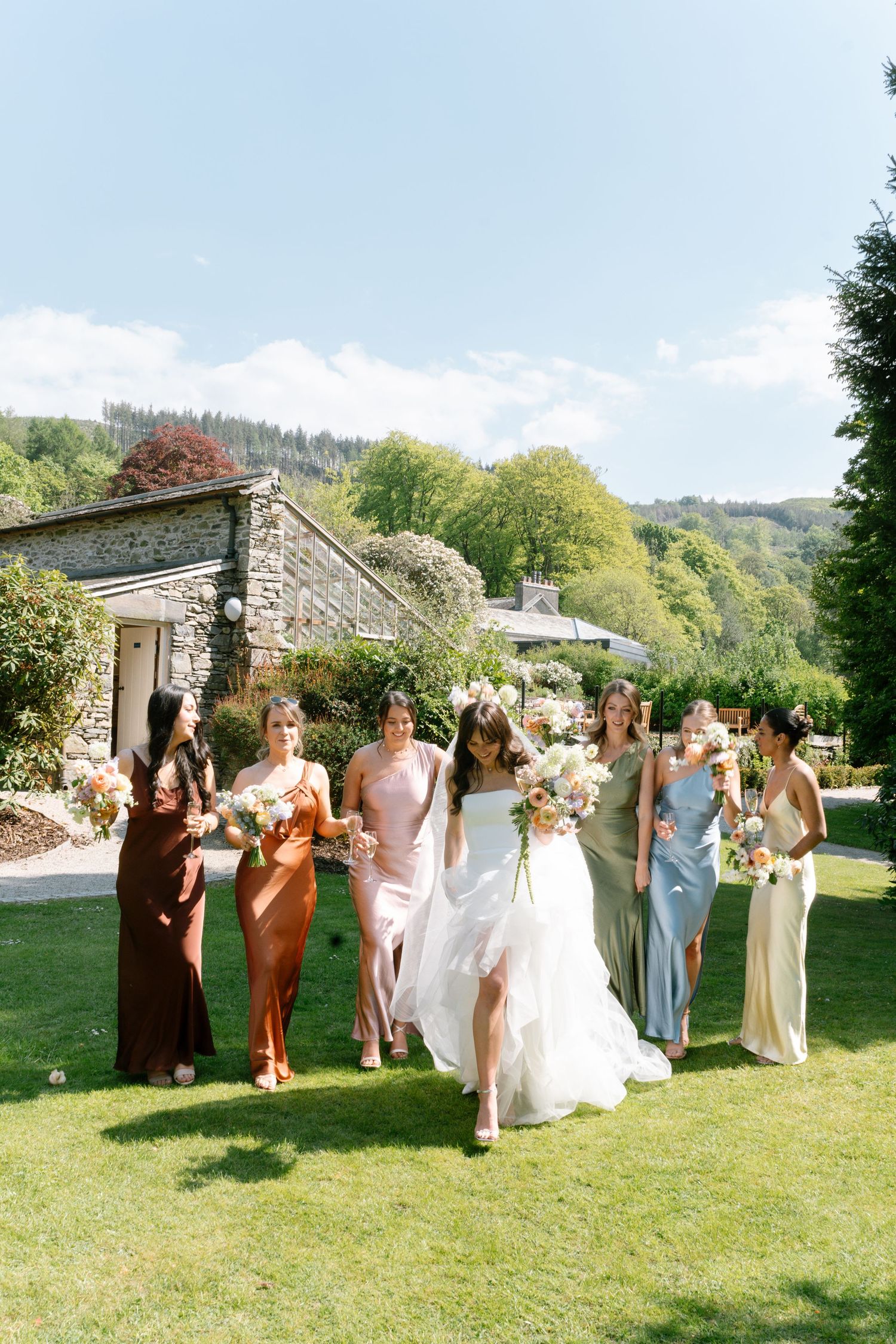 Wedding party in brown and pastel dresses poses together on a sunny lawn near a stone building.