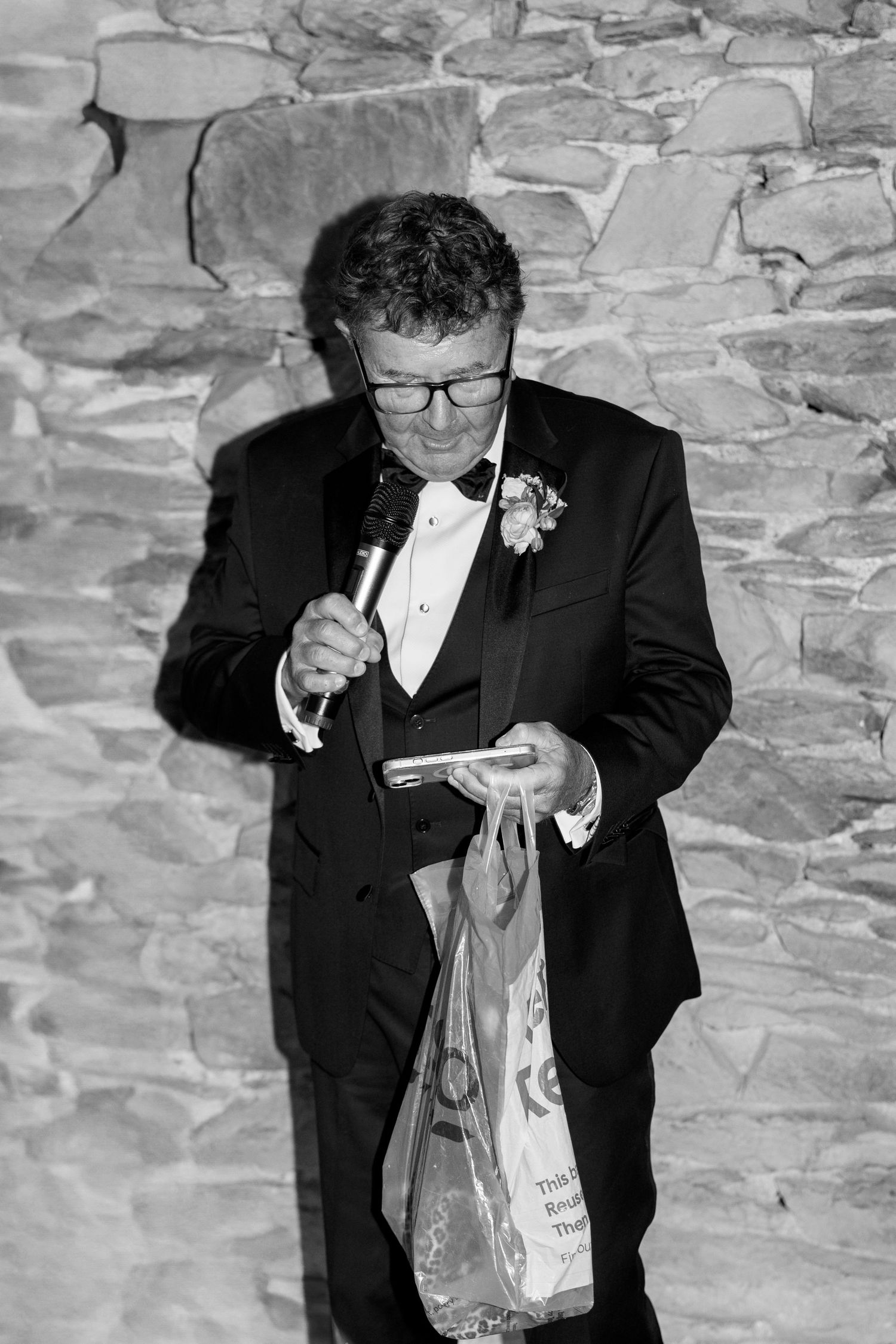 A person in a tuxedo reading from papers at a formal event against a stone wall background.