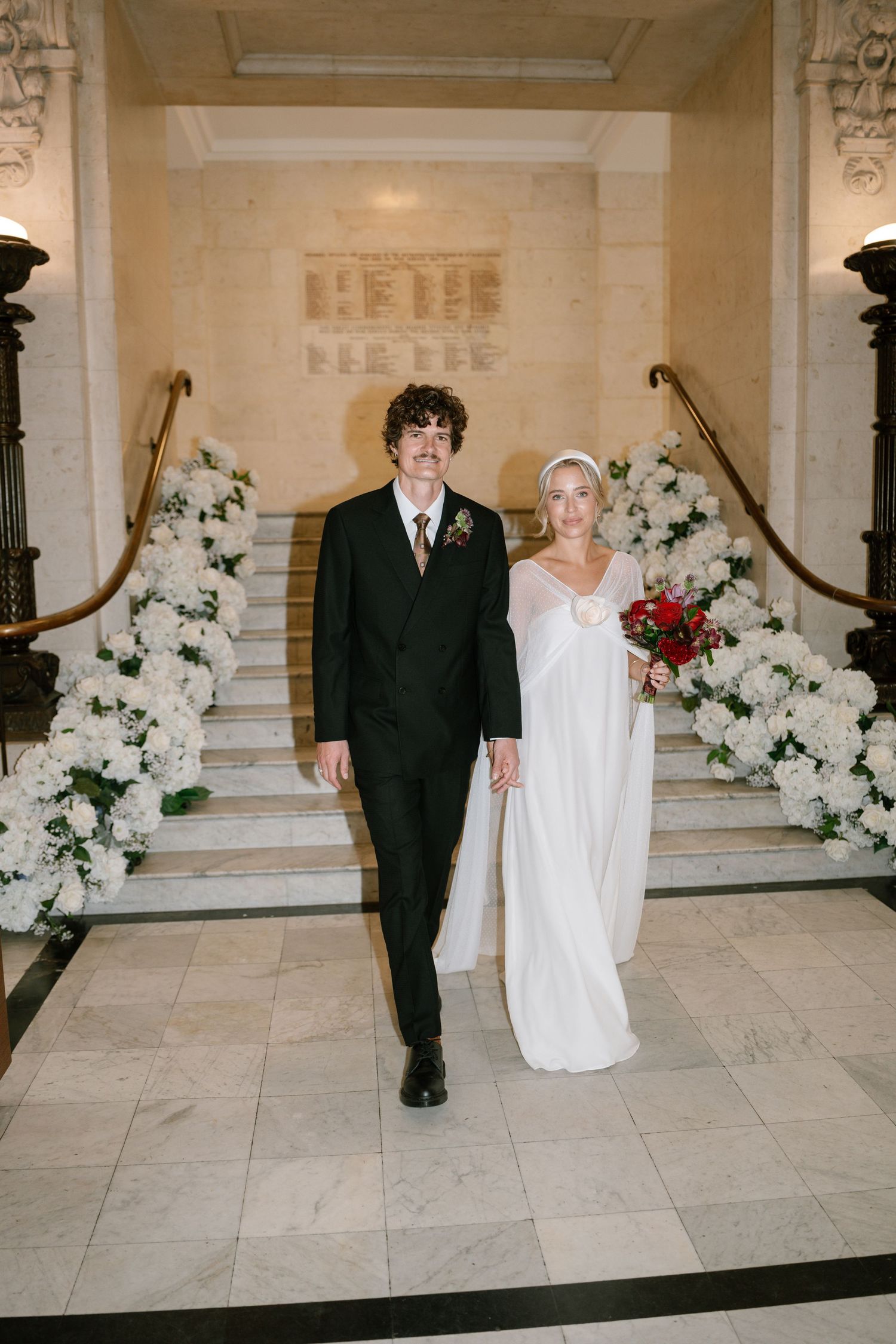 Wedding couple posing on marble stairs decorated with white floral arrangements.