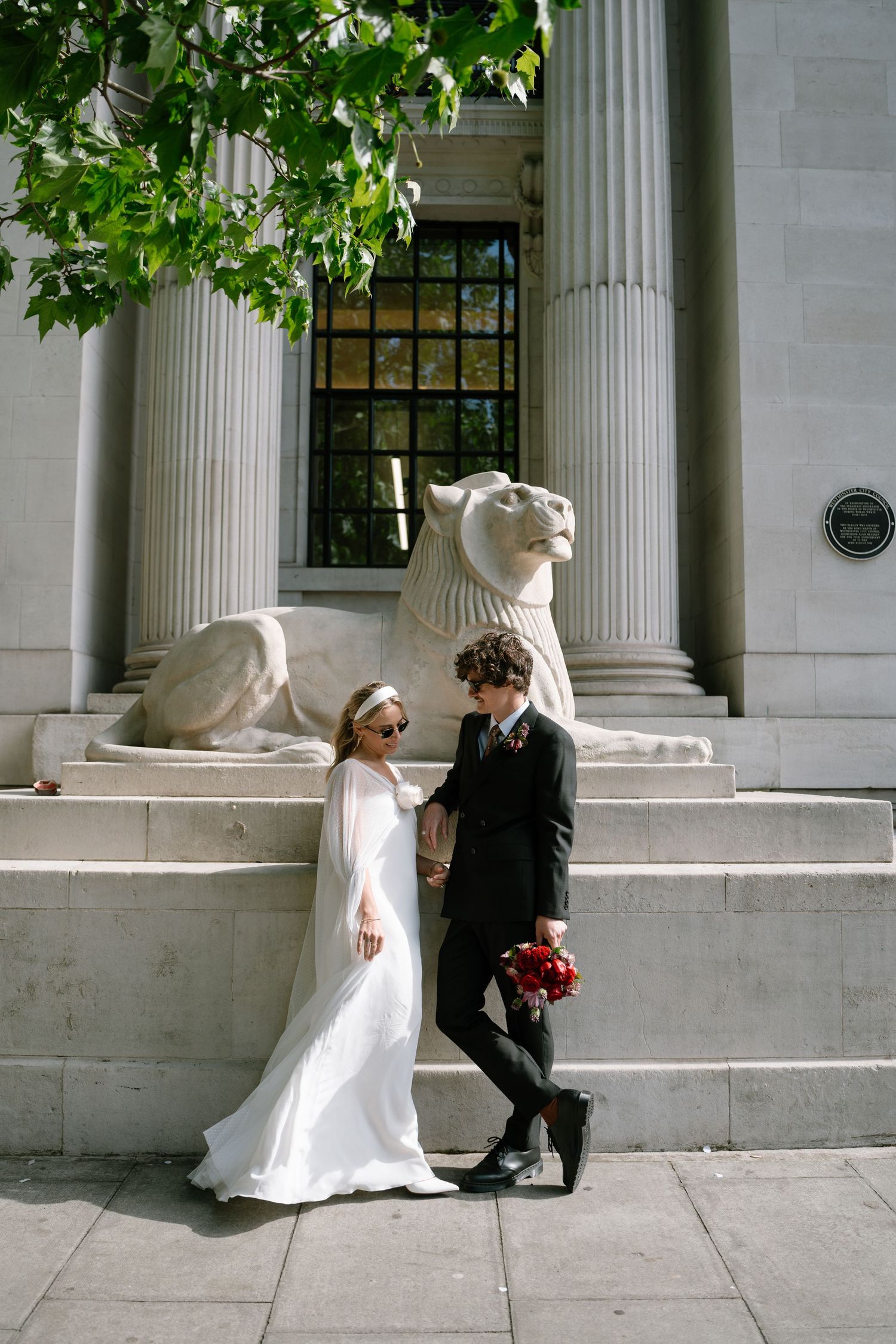A couple in wedding attire poses playfully by stone columns and lion statues.