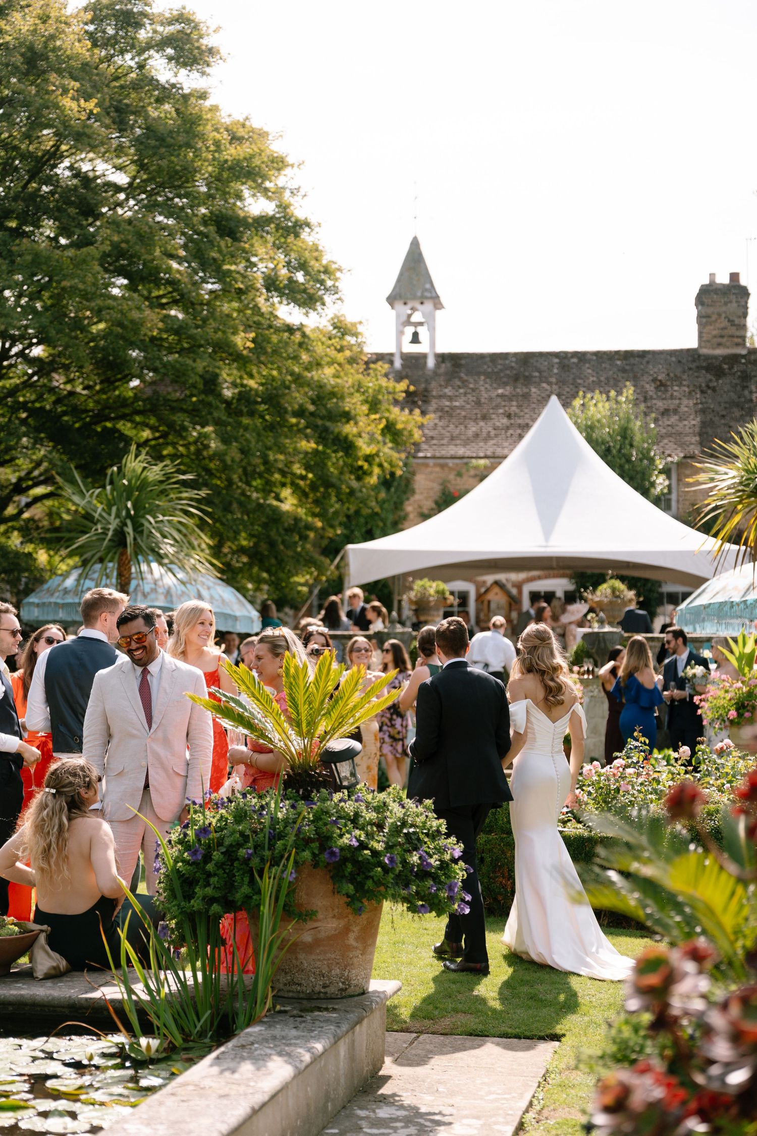 Wedding guests mingle at an outdoor reception with white tents and stone buildings in the background.