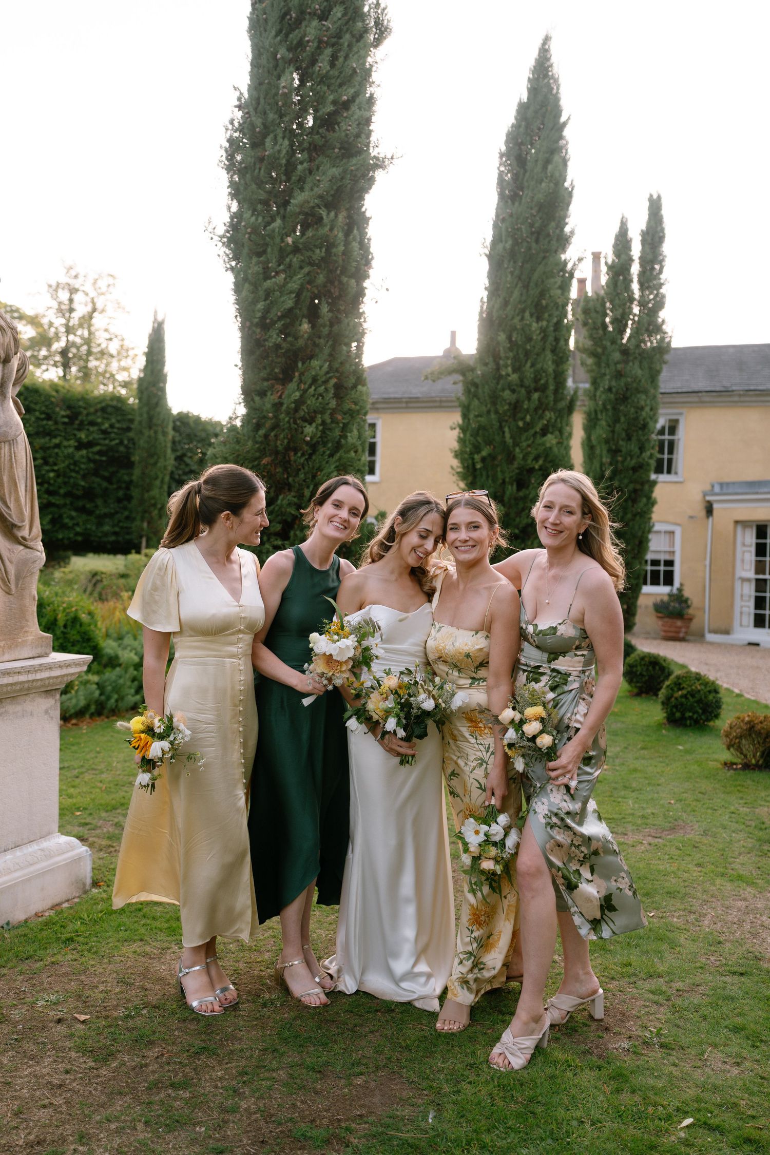 Bridesmaids in champagne dresses hold bouquets while gathered on grass near cypress trees.