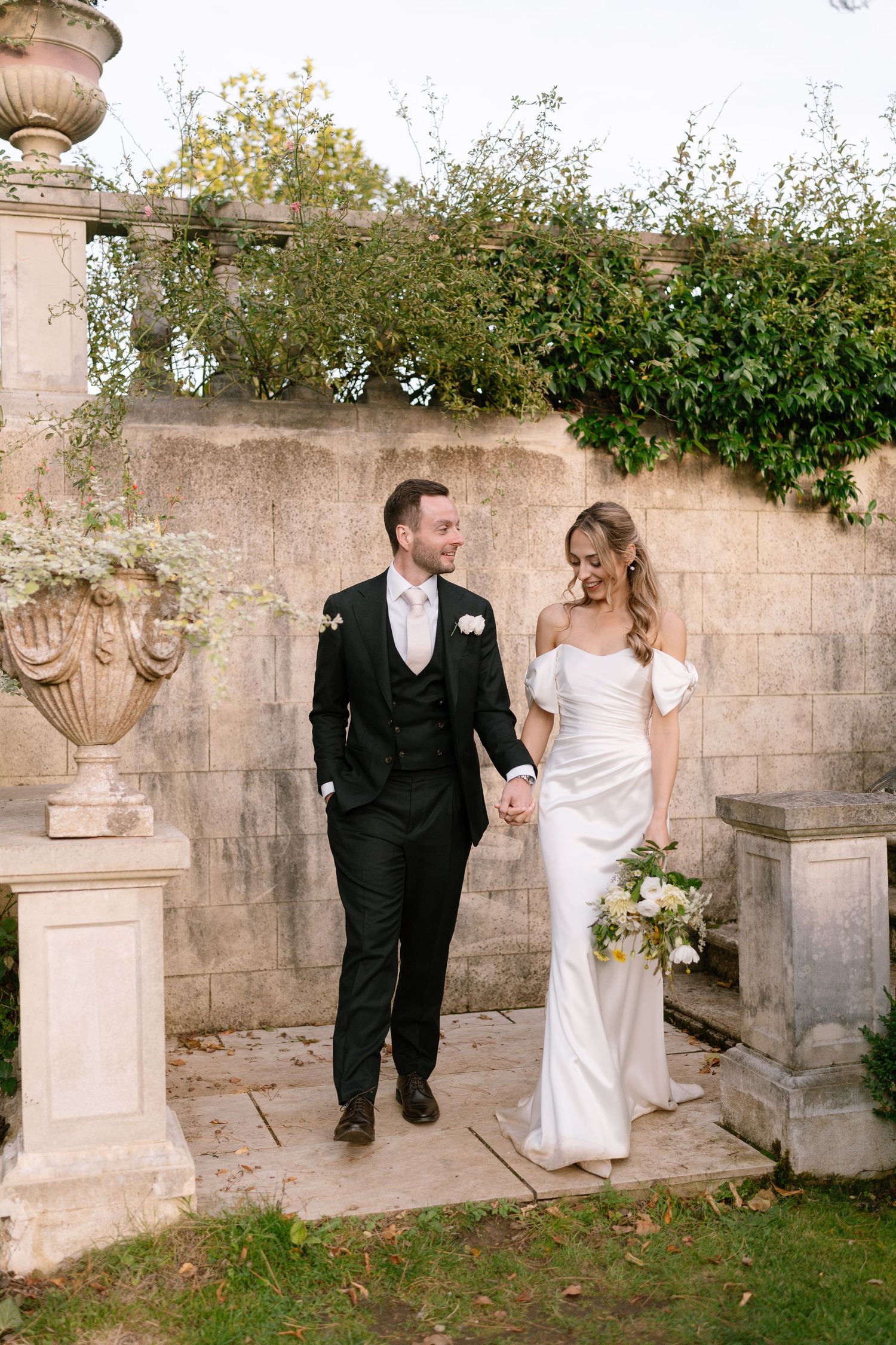 Couple walking along decorative stone wall with garden urns during wedding photos.