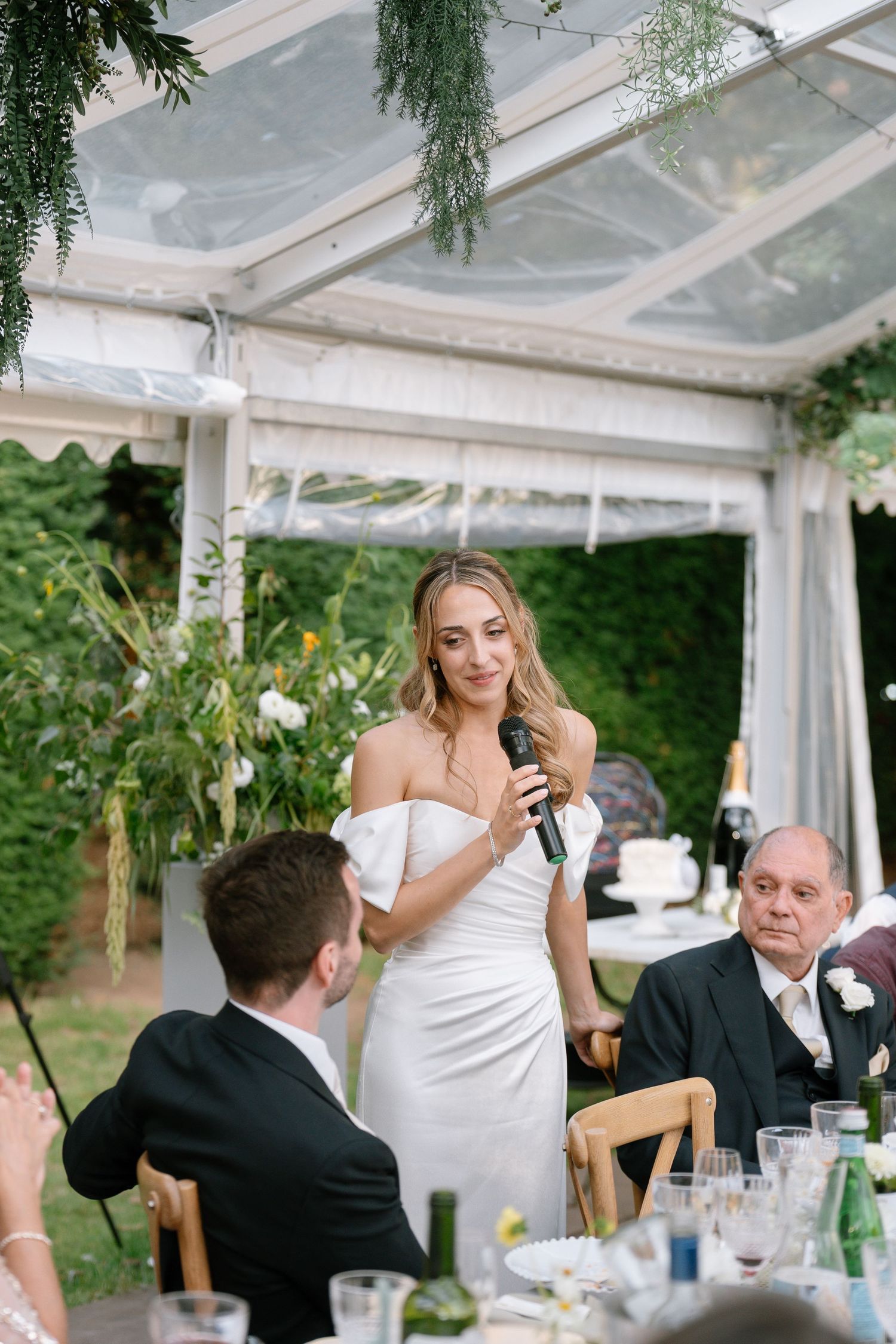 Wedding celebration in a clear tent with greenery decorations and wooden chairs.