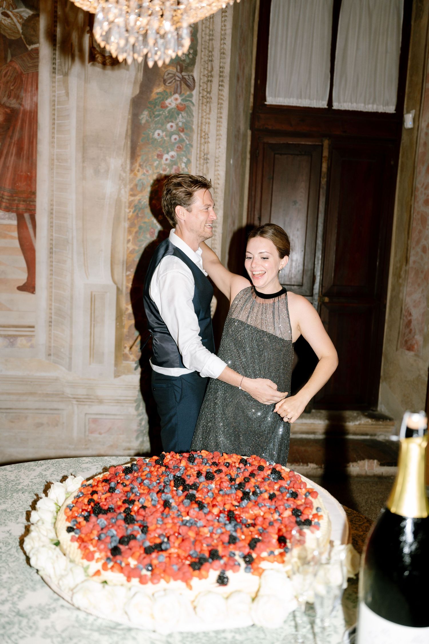 A couple celebrates near a large cake decorated with berries under an ornate chandelier.