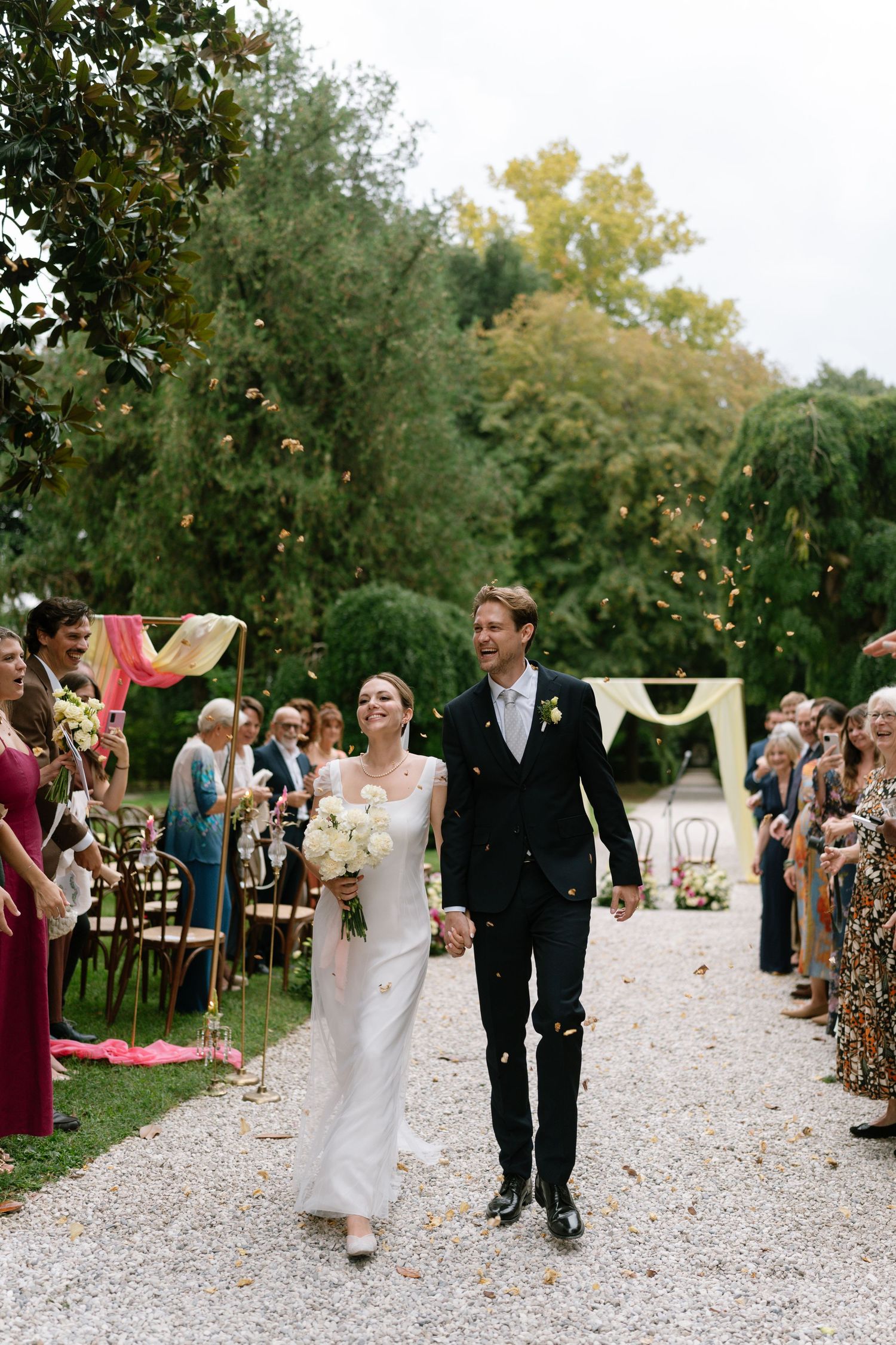 A joyful couple walks down a gravel aisle while wedding guests celebrate their union.