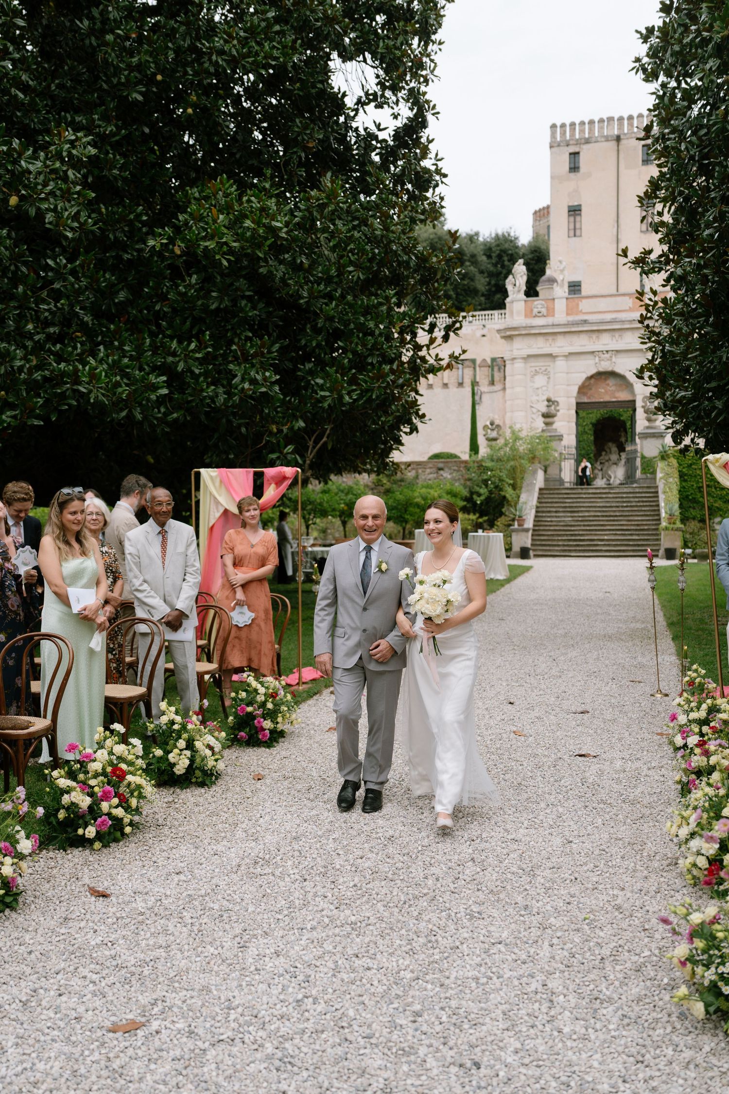 Wedding guests walk along a gravel path lined with floral arrangements leading to an elegant venue.