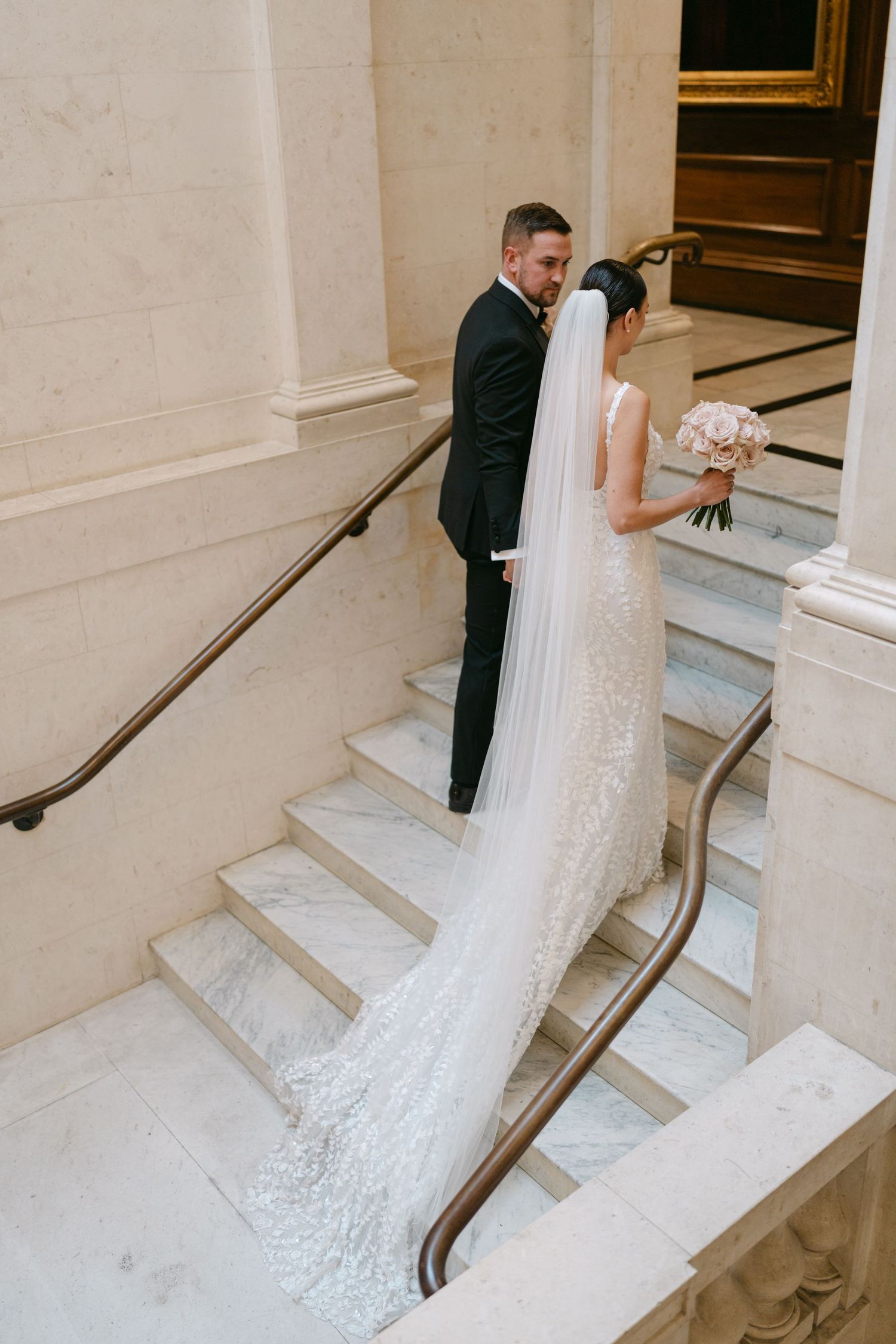 Couple poses on elegant marble staircase with bride's long veil cascading down steps.