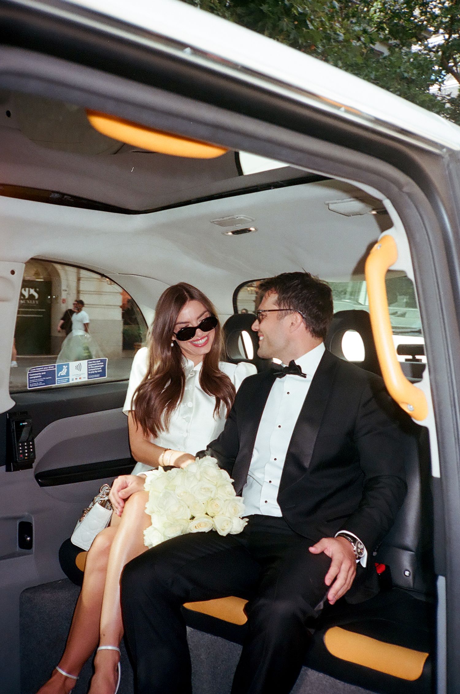 Bride and groom in wedding attire sitting in a London black cab, smiling at each other, bride holding white flowers.