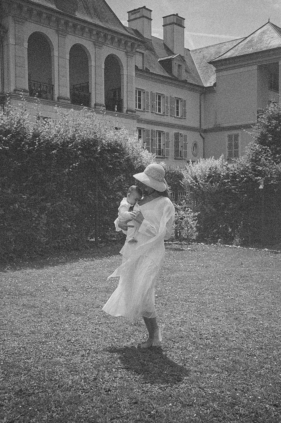 Black and white photo of a woman in white dress and hat holding a child in a grand estate garden.