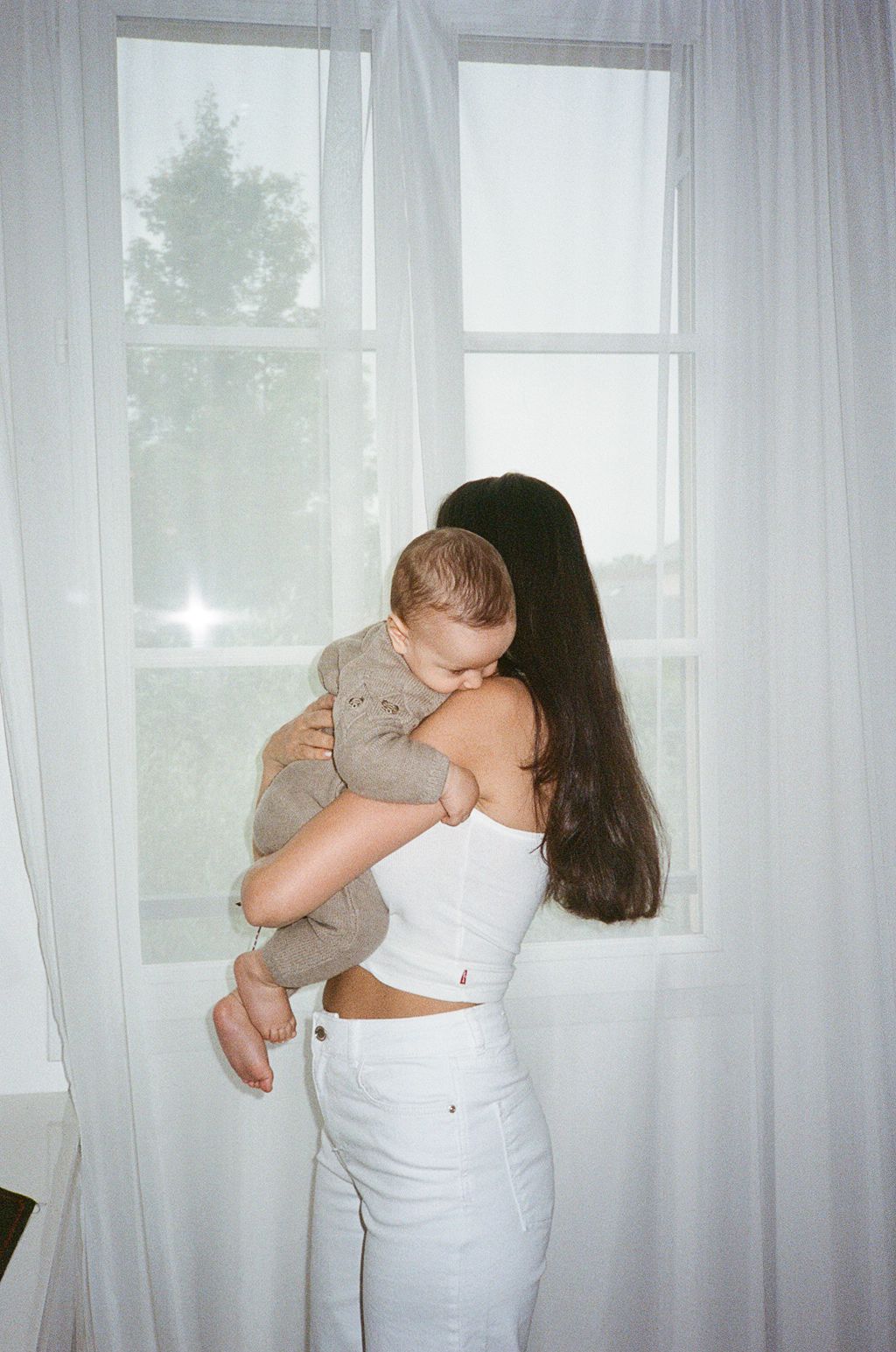 Young mother with long dark hair holding her baby near a bright window with white curtains indoors.