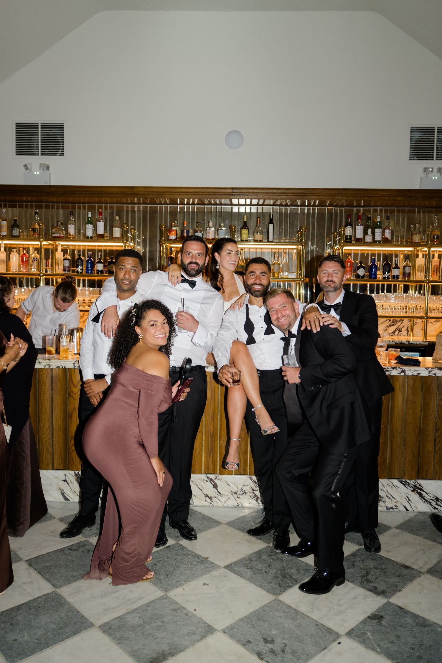 Festive wedding guests and bartenders posing together in front of an elegant marble and wood bar.