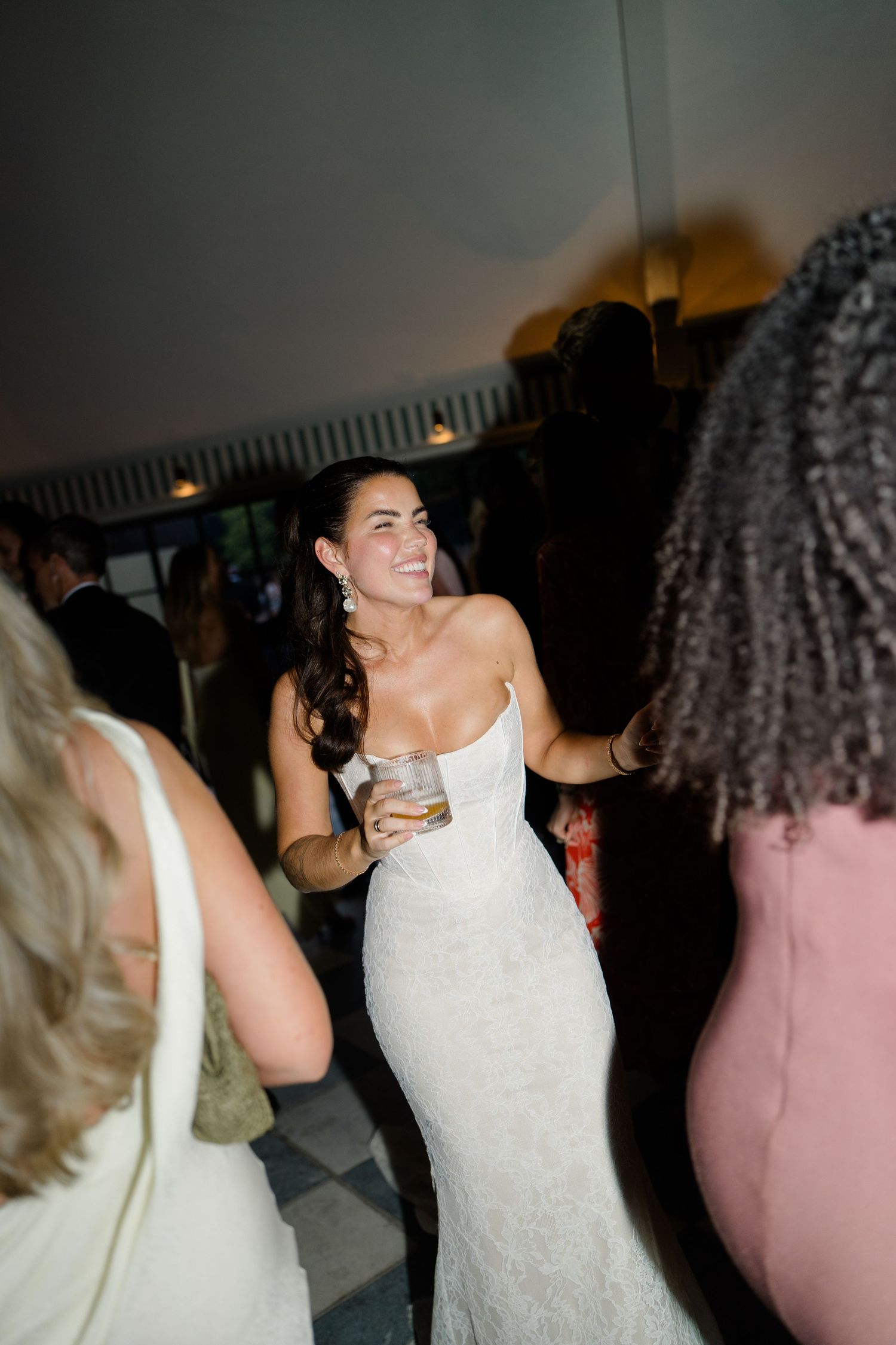 Radiant bride in strapless lace gown holds a drink while dancing at reception.