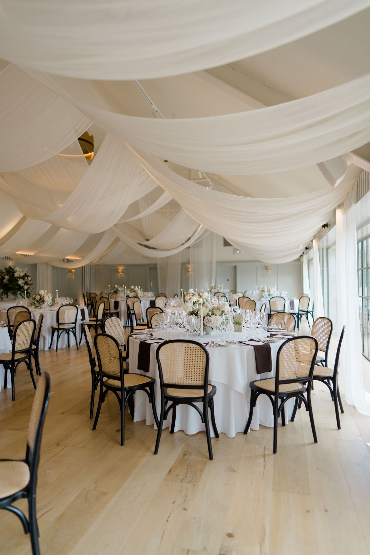Elegant wedding reception hall with white draped ceiling, bentwood chairs, and floral centerpieces.