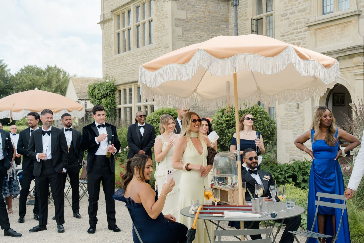 Wedding guests in formal attire gathered outside stone manor under a fringed peach parasol at reception.
