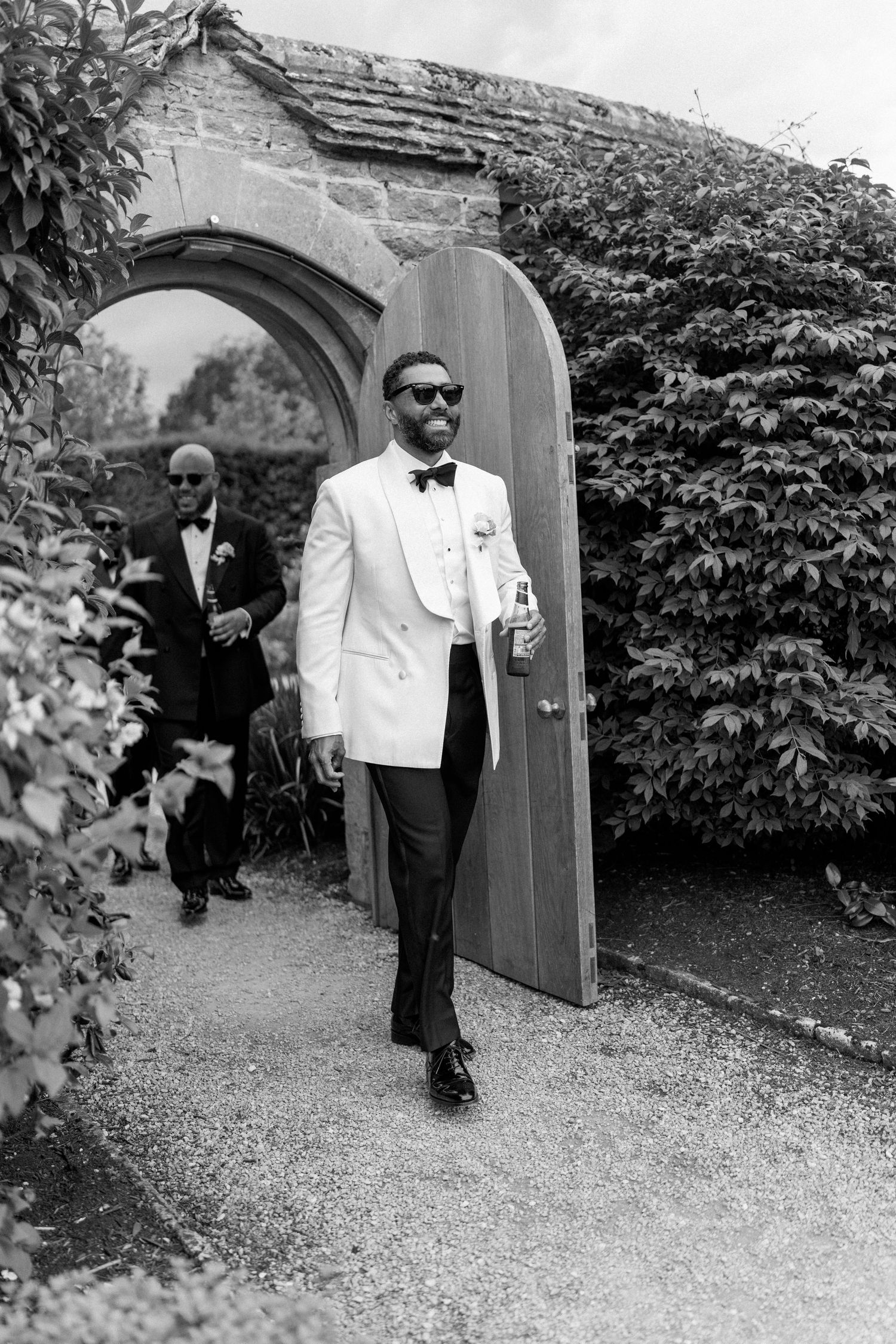 Stylish groom in white tuxedo jacket and sunglasses walks through garden arch at wedding venue.