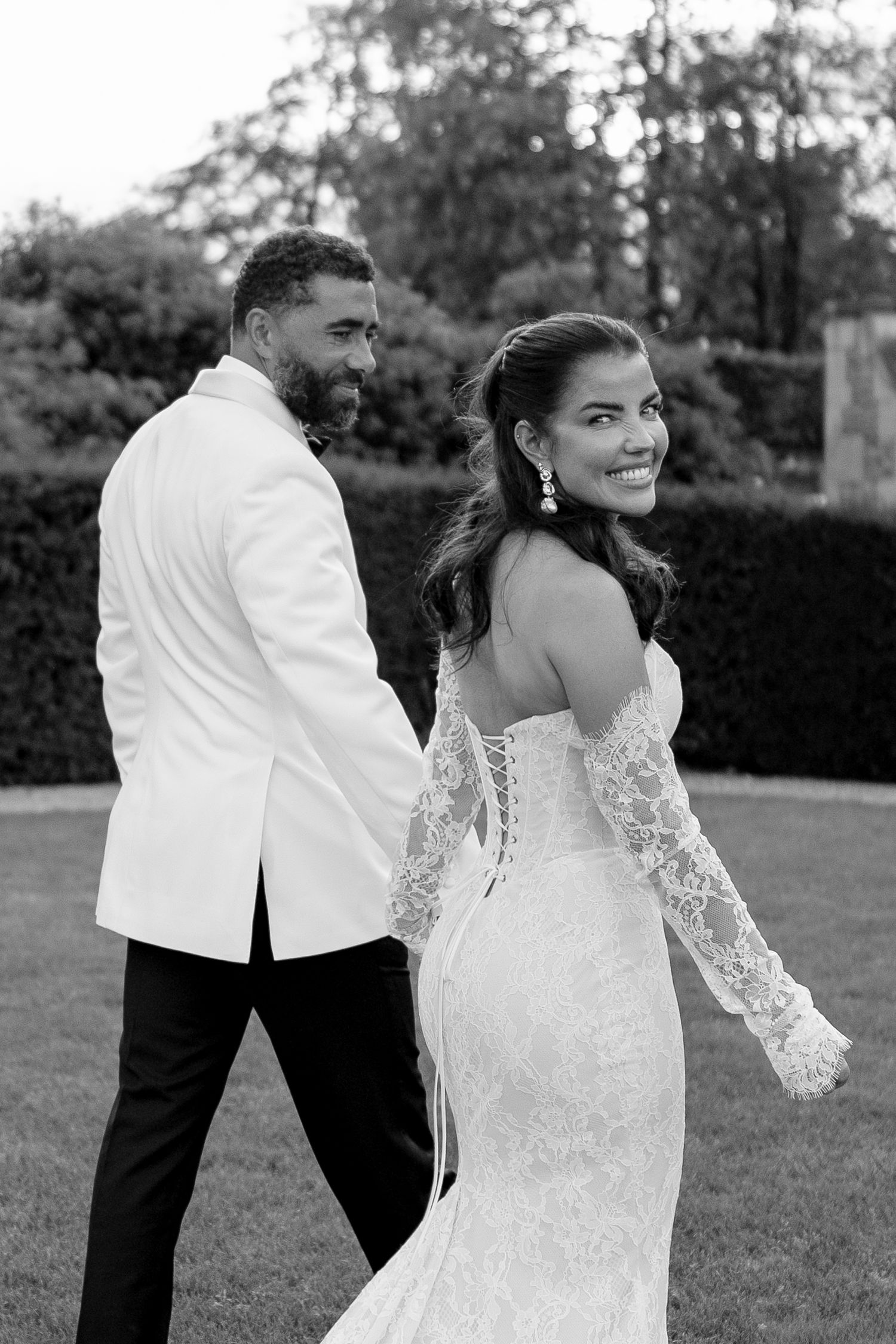 Black and white photo of bride in lace gown looking back smiling while walking with groom in white tuxedo.