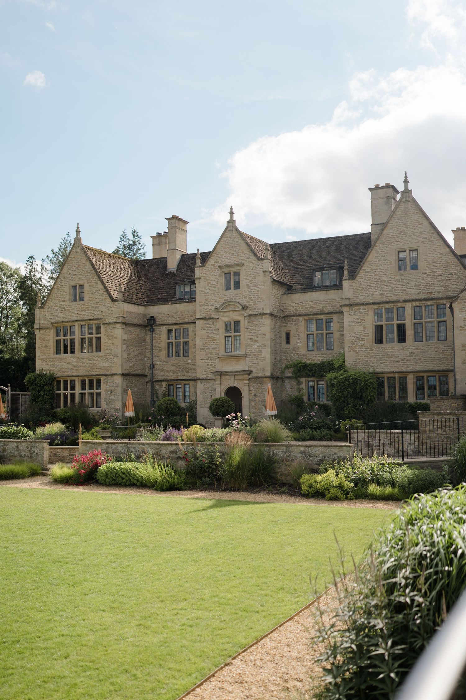 Elegant Cotswold stone manor house with manicured gardens and flowering borders under a blue sky.