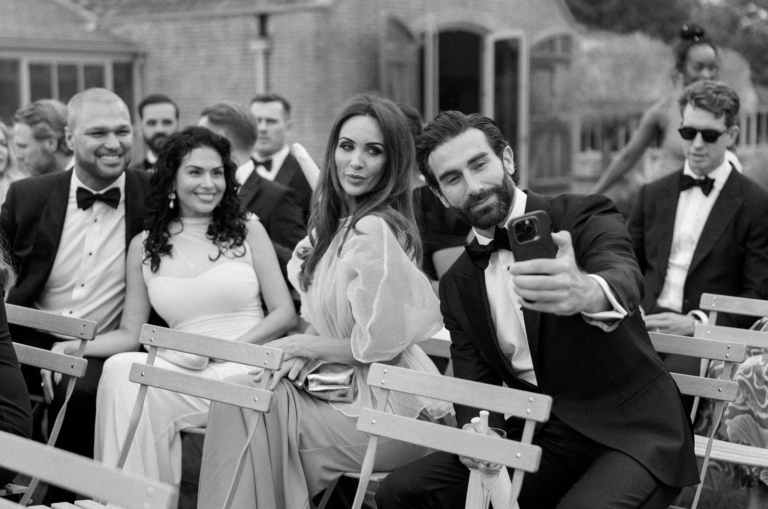 Black and white photo of wedding guests taking a selfie at an outdoor ceremony, dressed in formal attire.