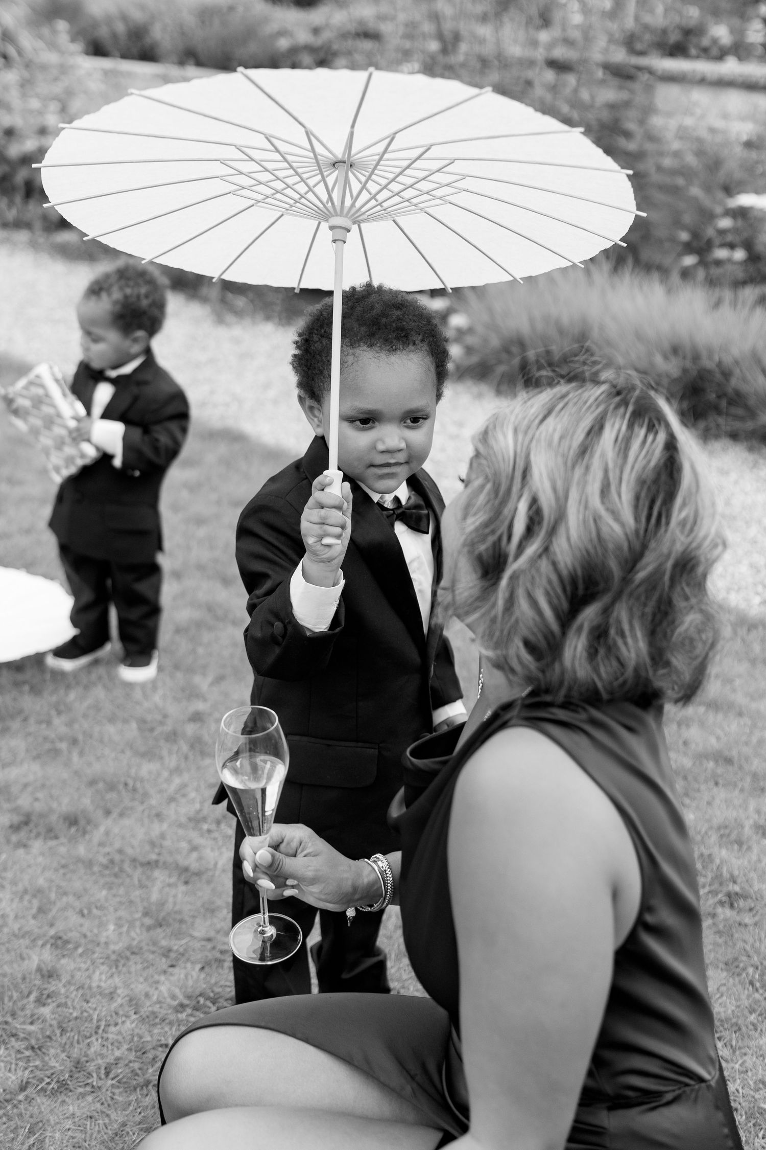 Adorable young boy in a tuxedo holding a white parasol at an outdoor wedding reception.