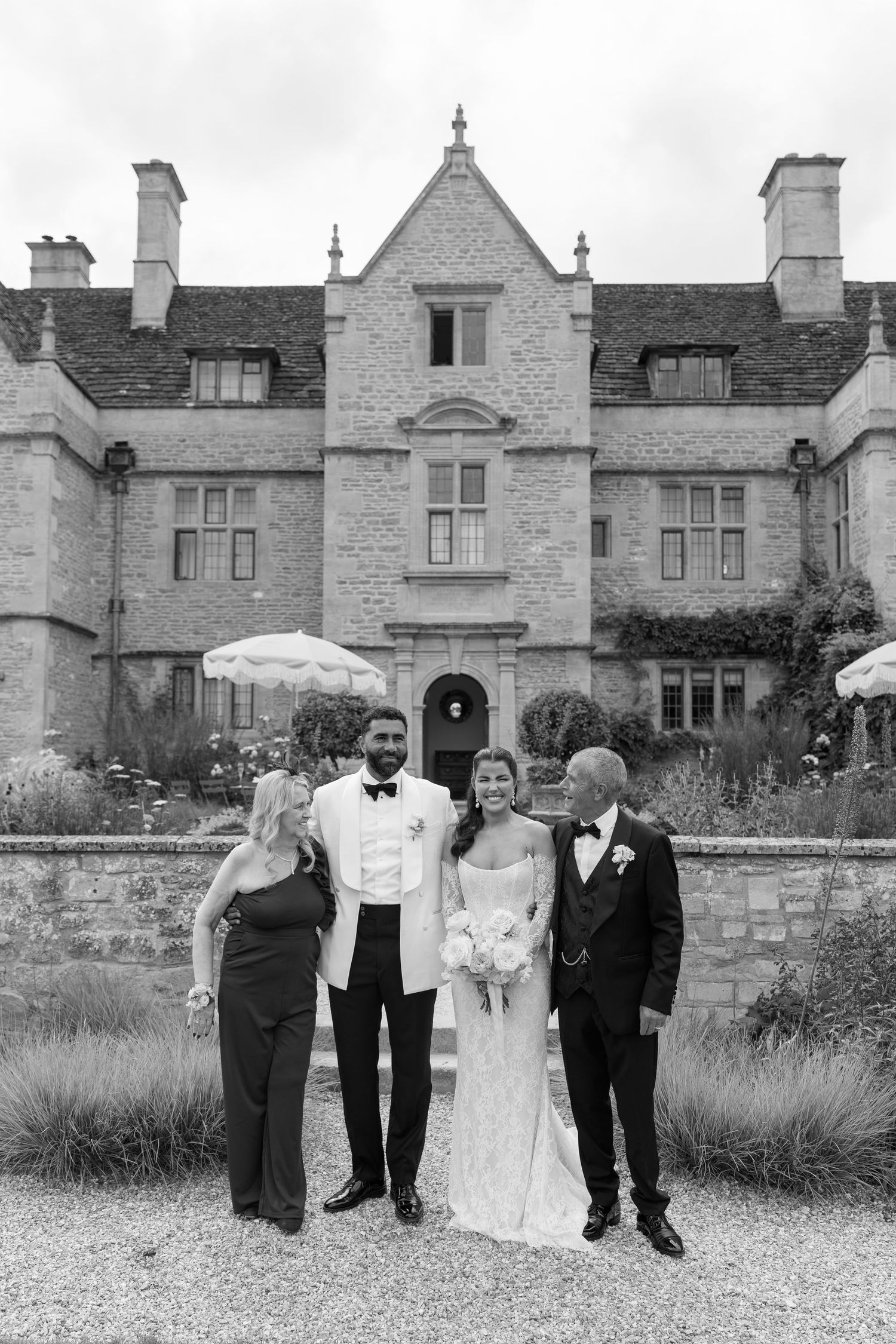 Black and white portrait of newlyweds and family posing before a historic stone manor house.