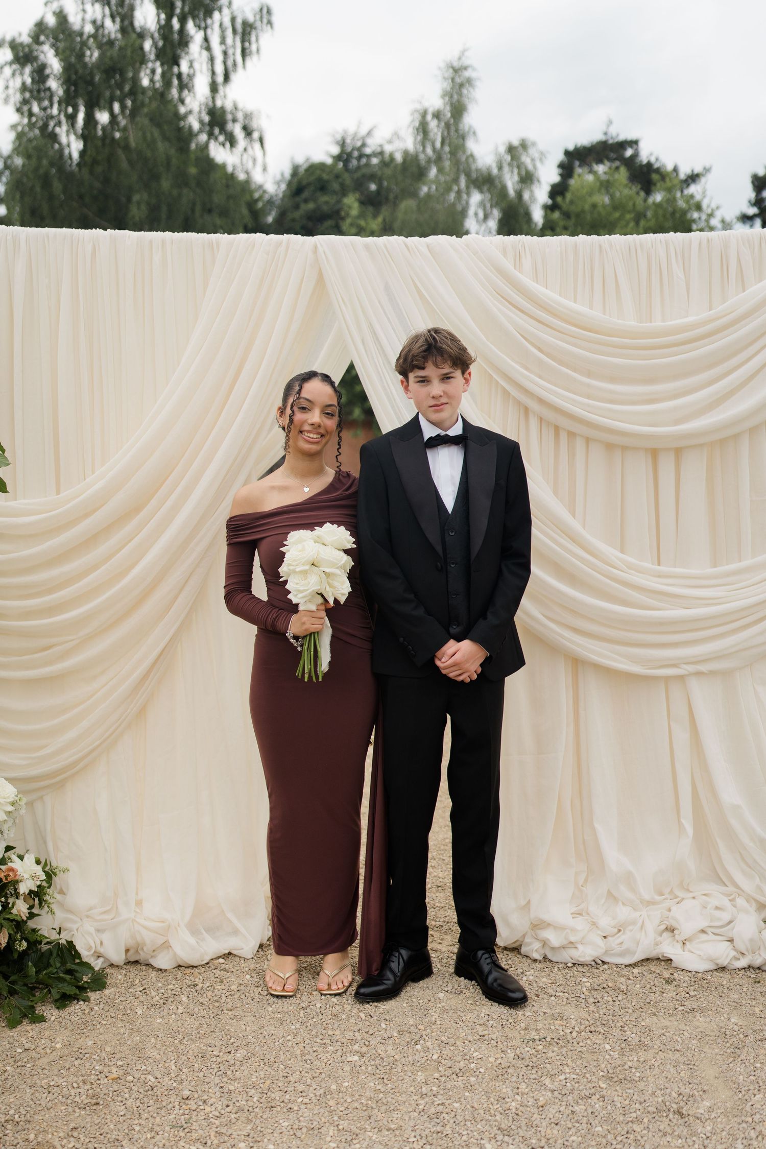 Bridesmaid in burgundy gown holding white bouquet posing with young man in tuxedo at draped backdrop.