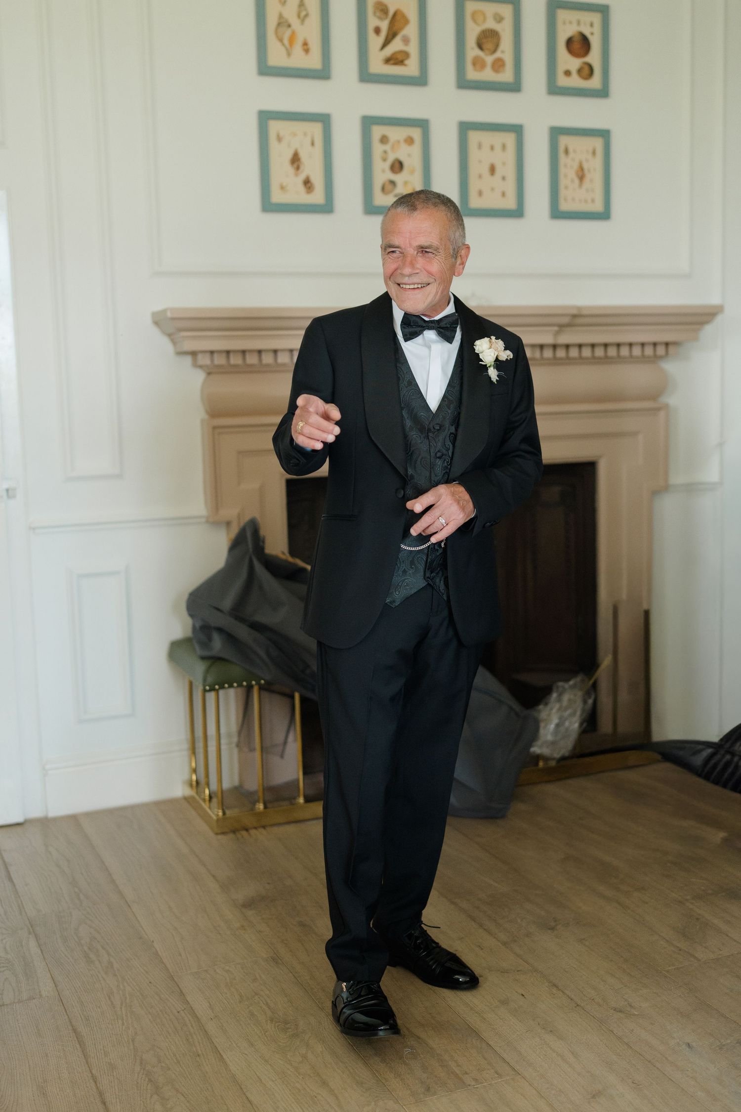 Smiling groom in black tuxedo with bow tie and boutonniere poses by fireplace in elegant room.