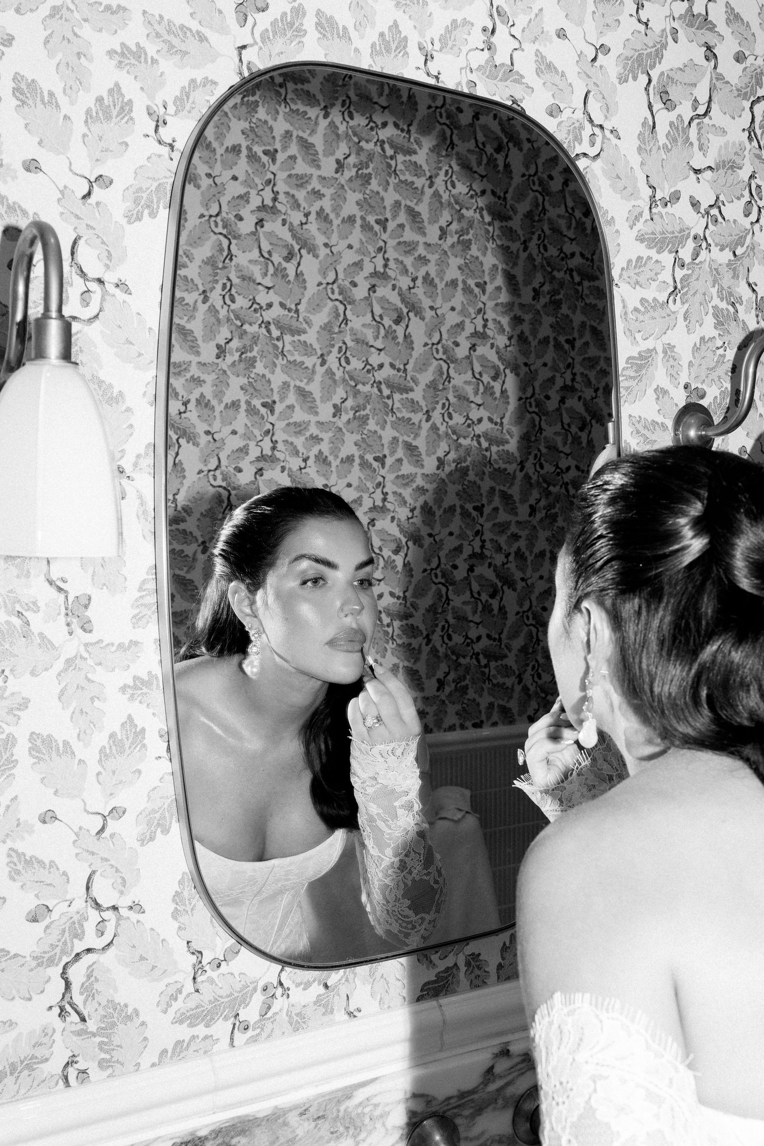 Black and white photo of a bride applying lipstick while looking into an ornate mirror with floral wallpaper.
