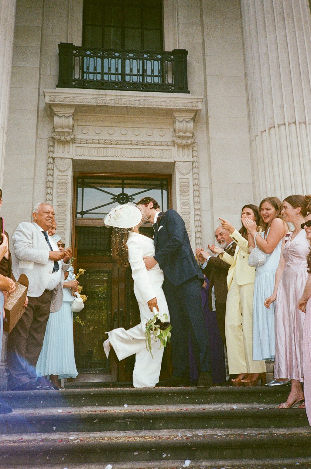Newlywed couple sharing a kiss on steps of elegant building, surrounded by applauding wedding guests in formal attire.