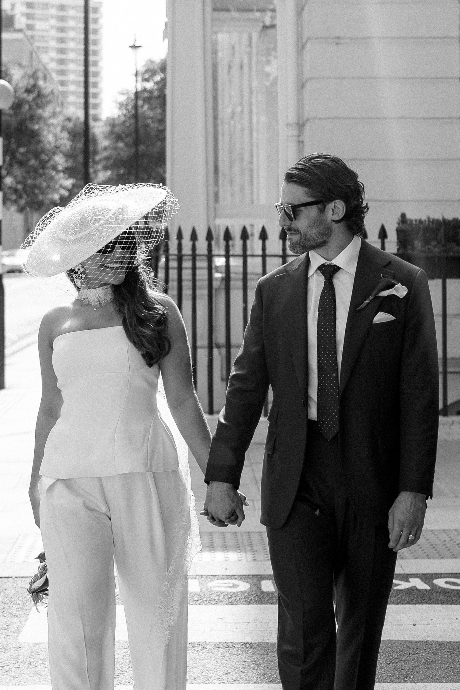Black and white photo of a bride in white jumpsuit and veil hat holding hands with groom in suit on city street.