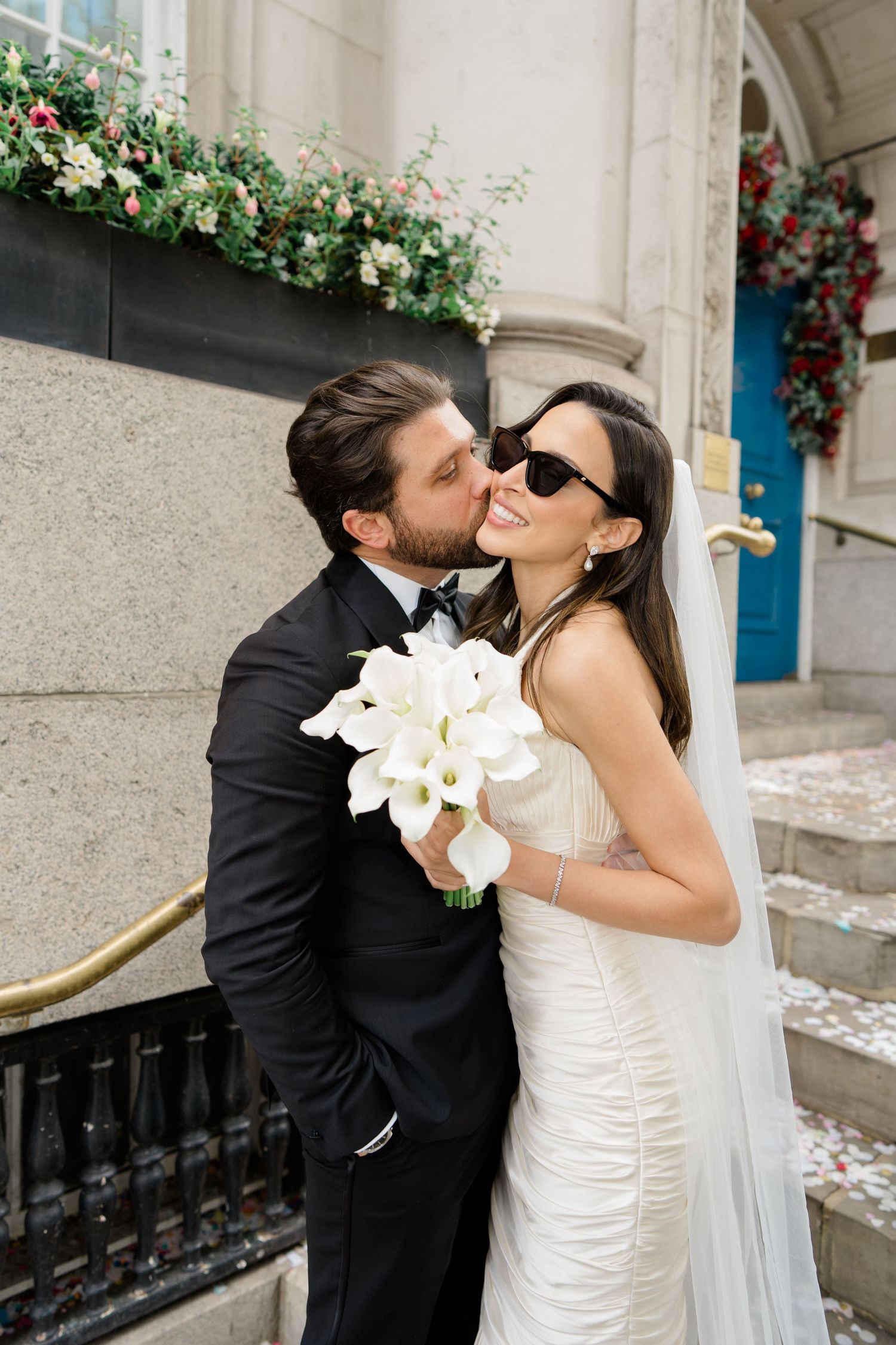 Groom in black tuxedo kissing bride wearing white dress, veil, and sunglasses holding white calla lily bouquet outdoors.