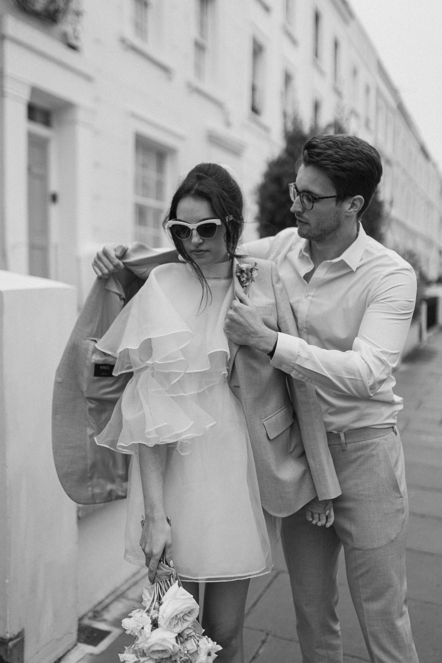 Black and white photo of a groom placing his jacket over a bride in a short ruffled dress and sunglasses holding a bouquet.